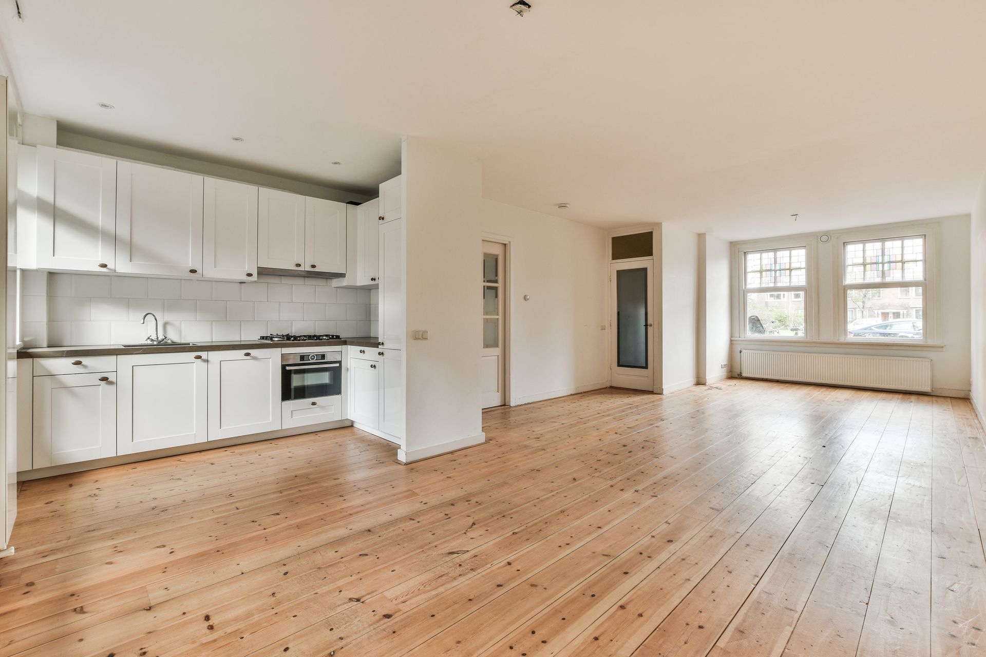 Empty room with white kitchen cabinets, wooden floor, and windows.