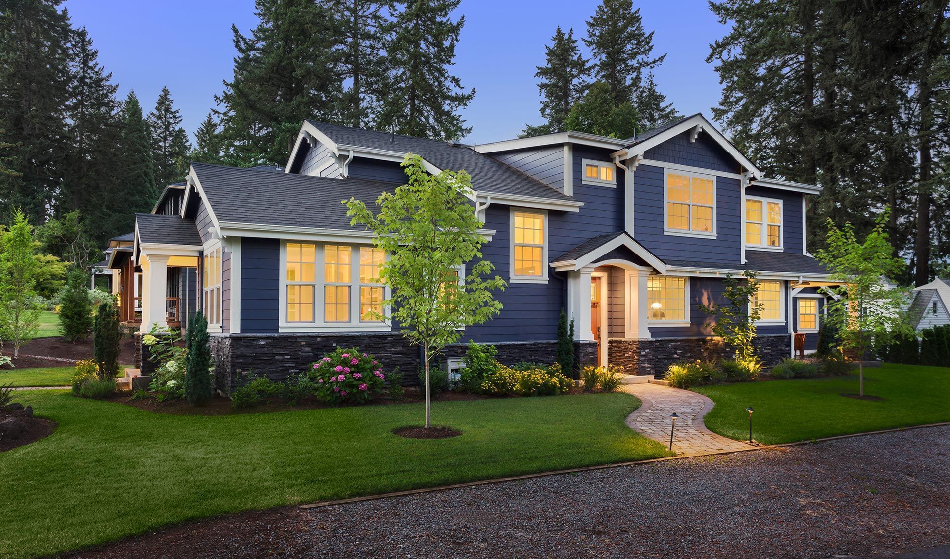 Dark blue two-story house with illuminated windows at dusk, stone path, green lawn, trees.