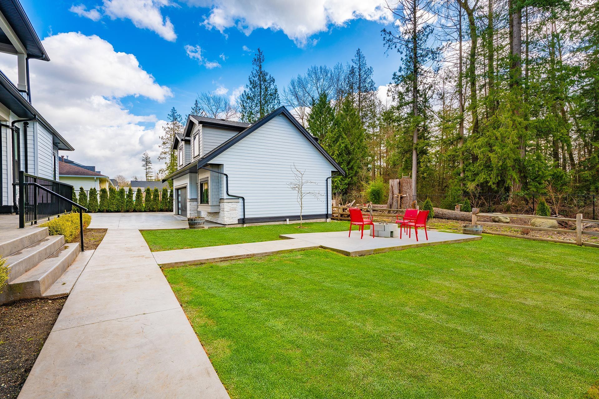 Lawn with pathway, red chairs around a fire pit, next to a garage and forest under a blue sky.