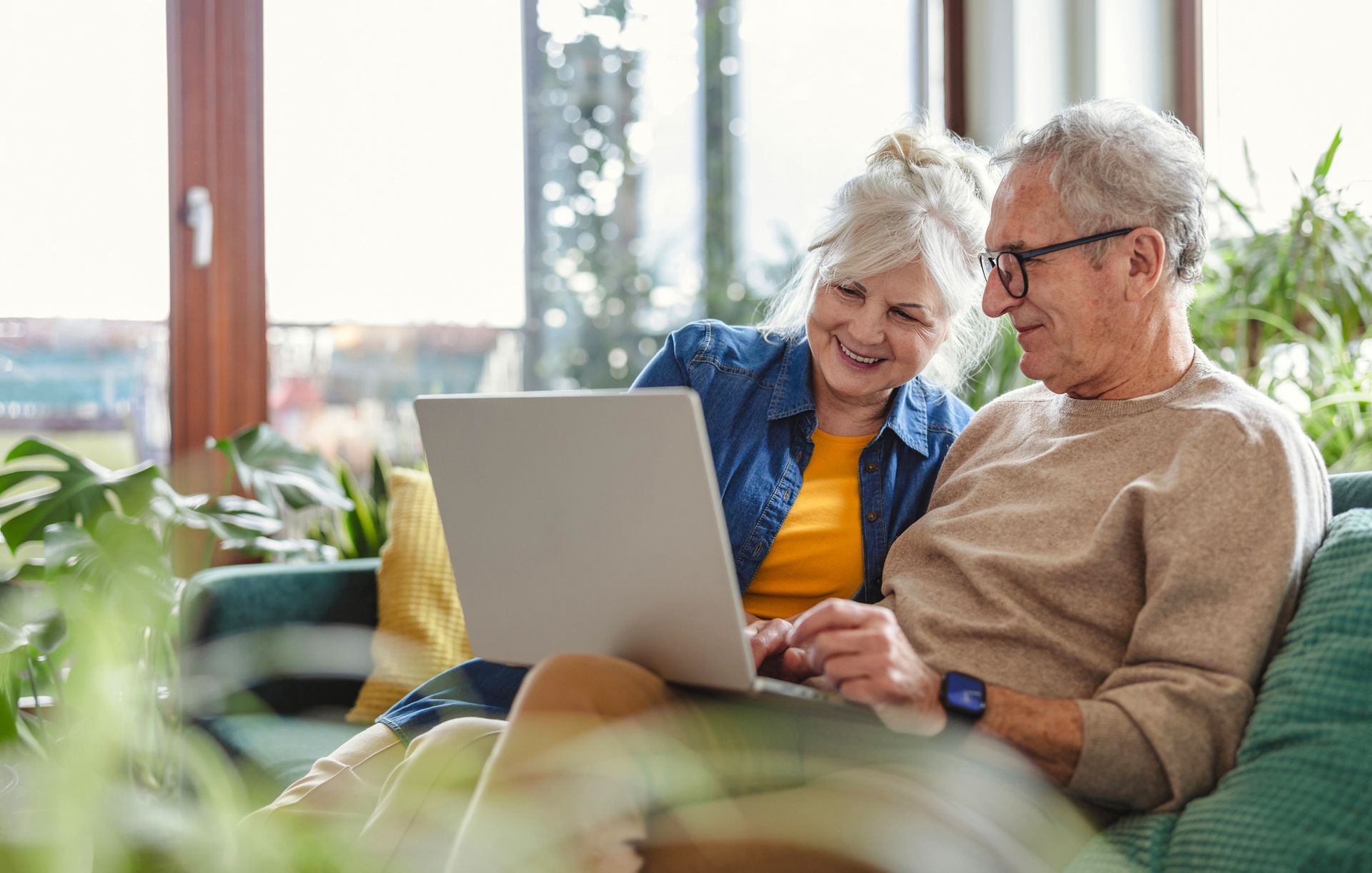 An elderly couple is sitting on a couch looking at a laptop computer.
