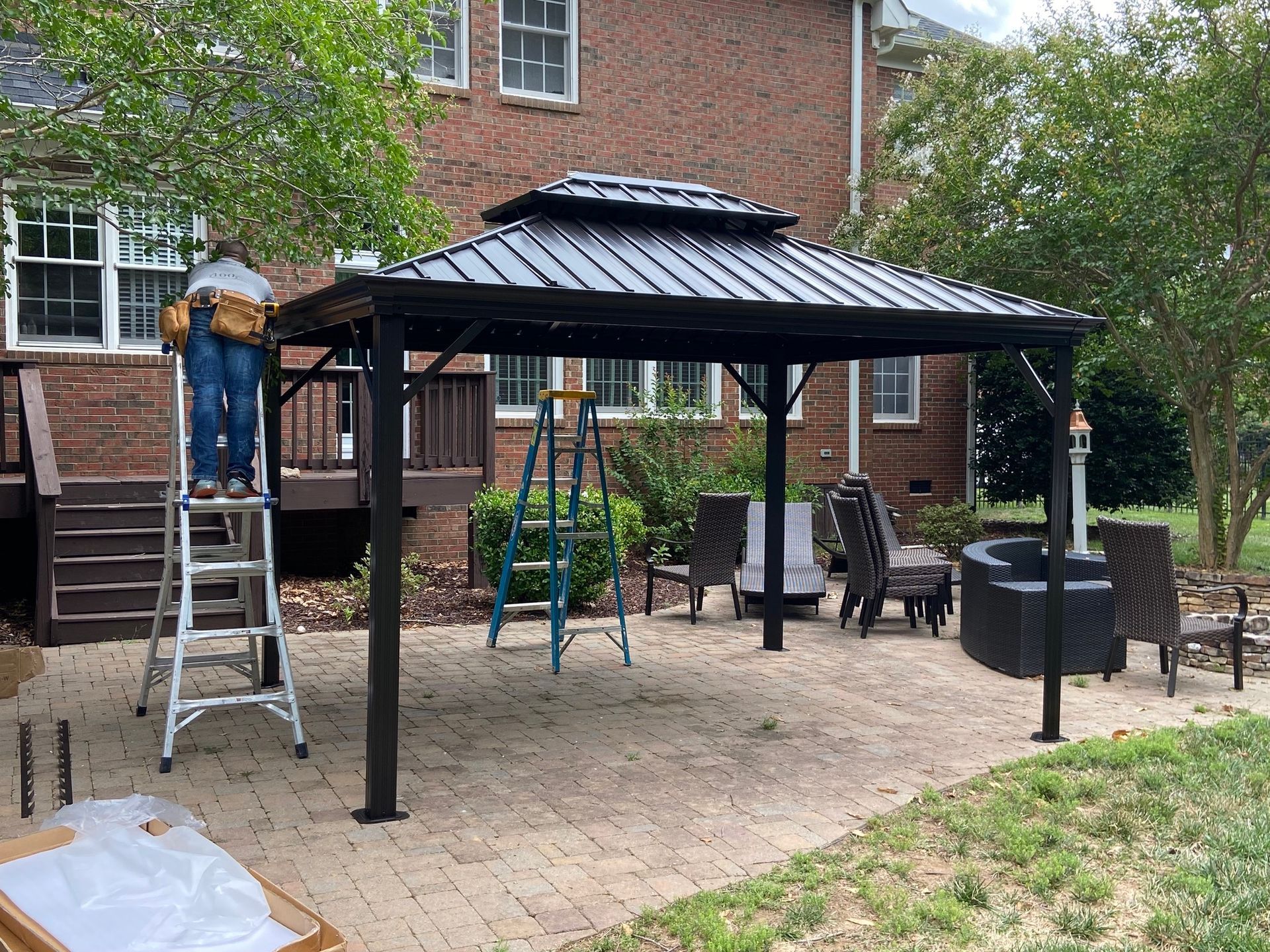 Person on a ladder assembling a black gazebo on a brick patio, with a house in the background.