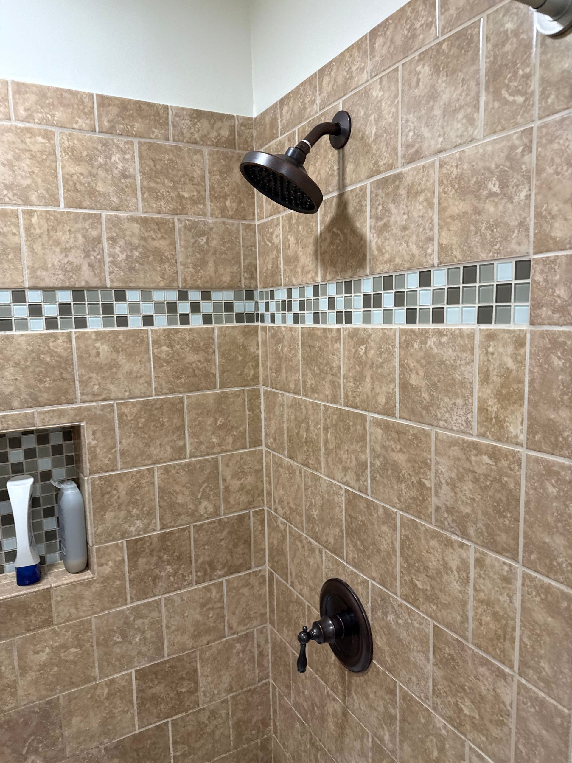 A tiled shower with a bronze showerhead, fixtures, and a mosaic tile border. A built-in shelf holds toiletries.