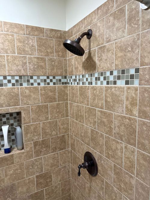 Tiled shower with a dark bronze showerhead and fixtures, plus a decorative mosaic tile band. A built-in shelf holds toiletries.