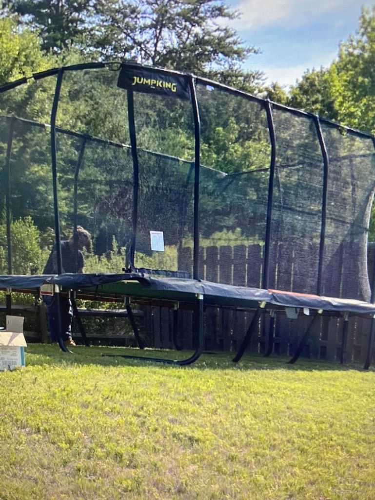 Black trampoline with safety net on green grass, with a wooden fence and trees in the background.
