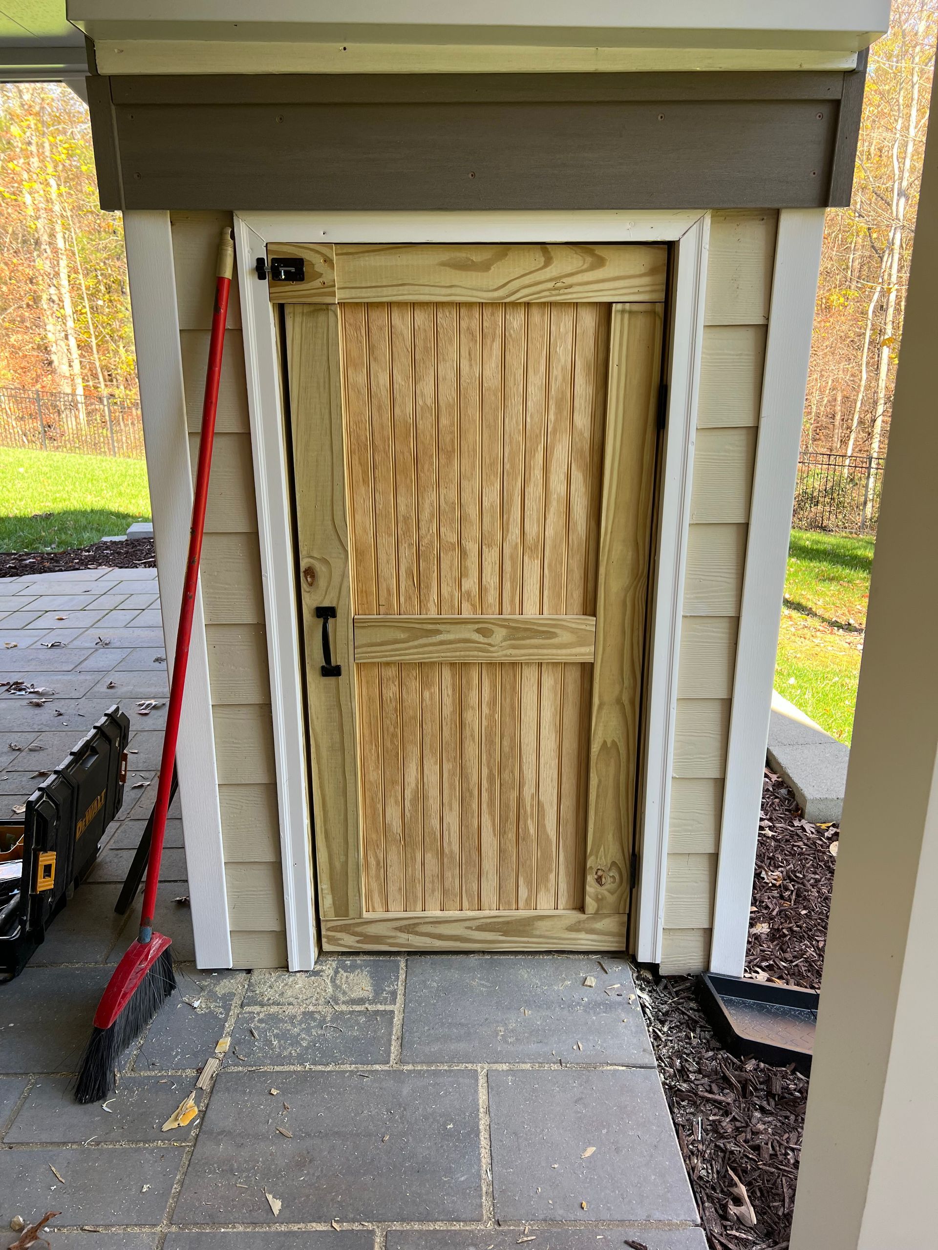 Wooden door in a recessed alcove with light siding and a stone floor. A broom and tools are nearby.