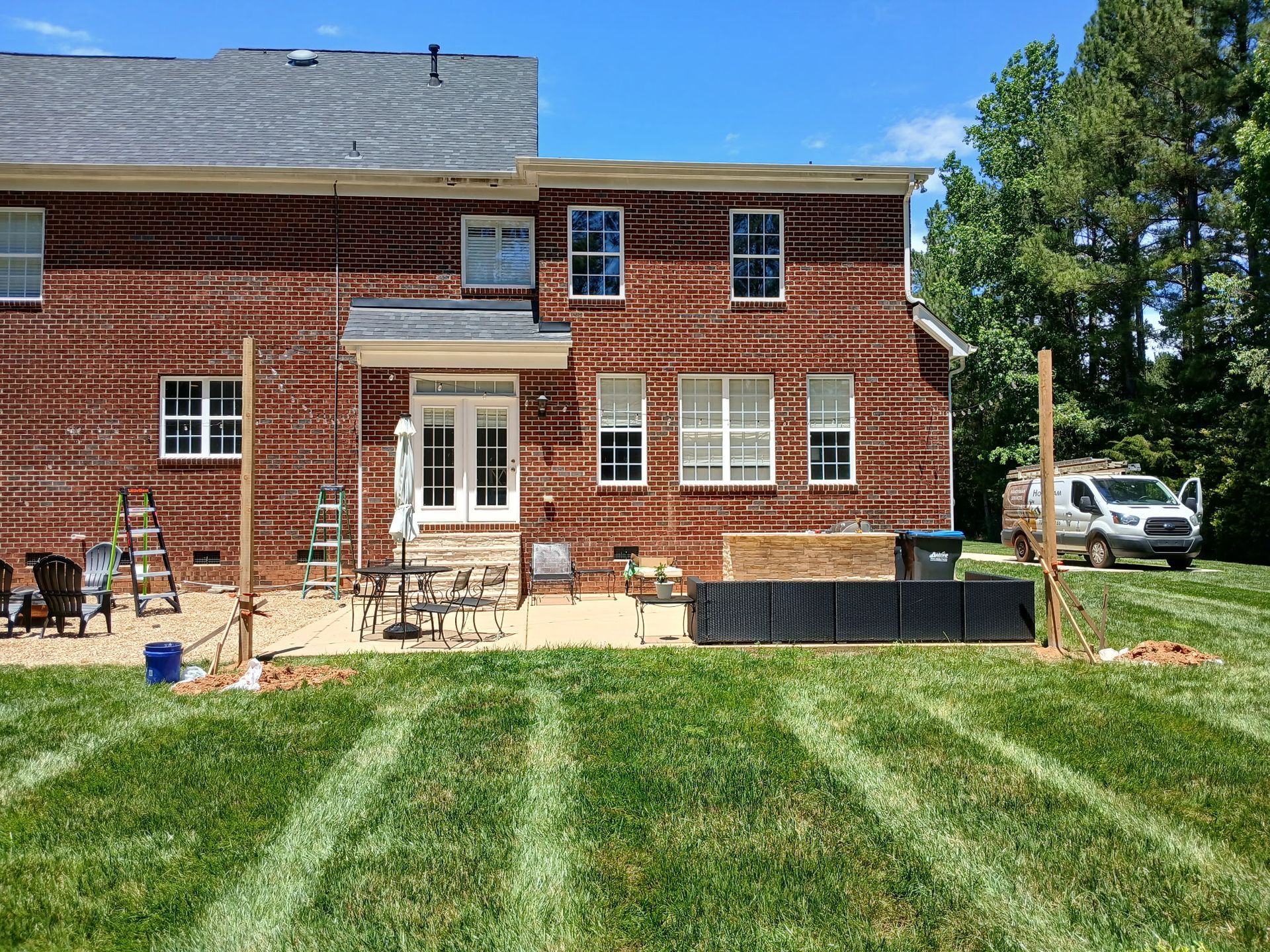 A brick house with a patio, lawn, and parked van. Wooden posts are set up in the grass, possibly for construction.