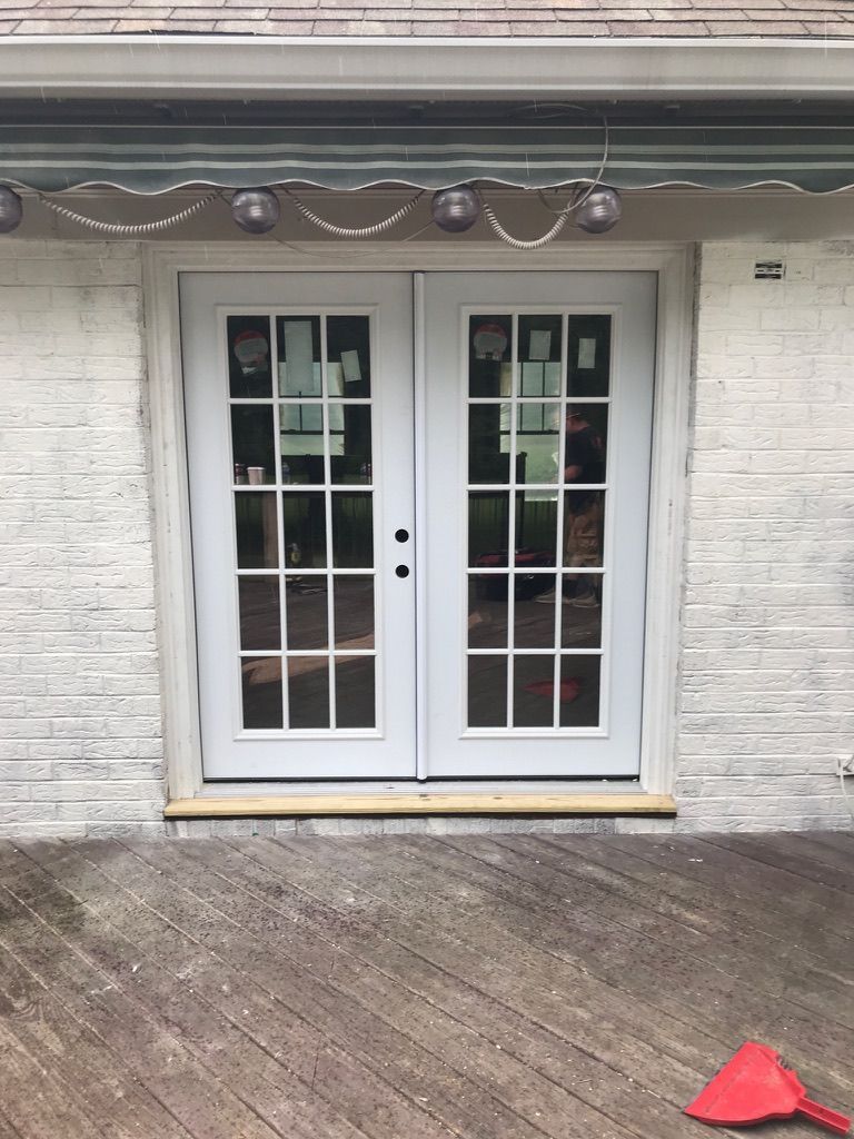 White double doors with glass panes, set in a textured white brick wall. A wooden deck is in front of the doors; a red object is in the lower right.