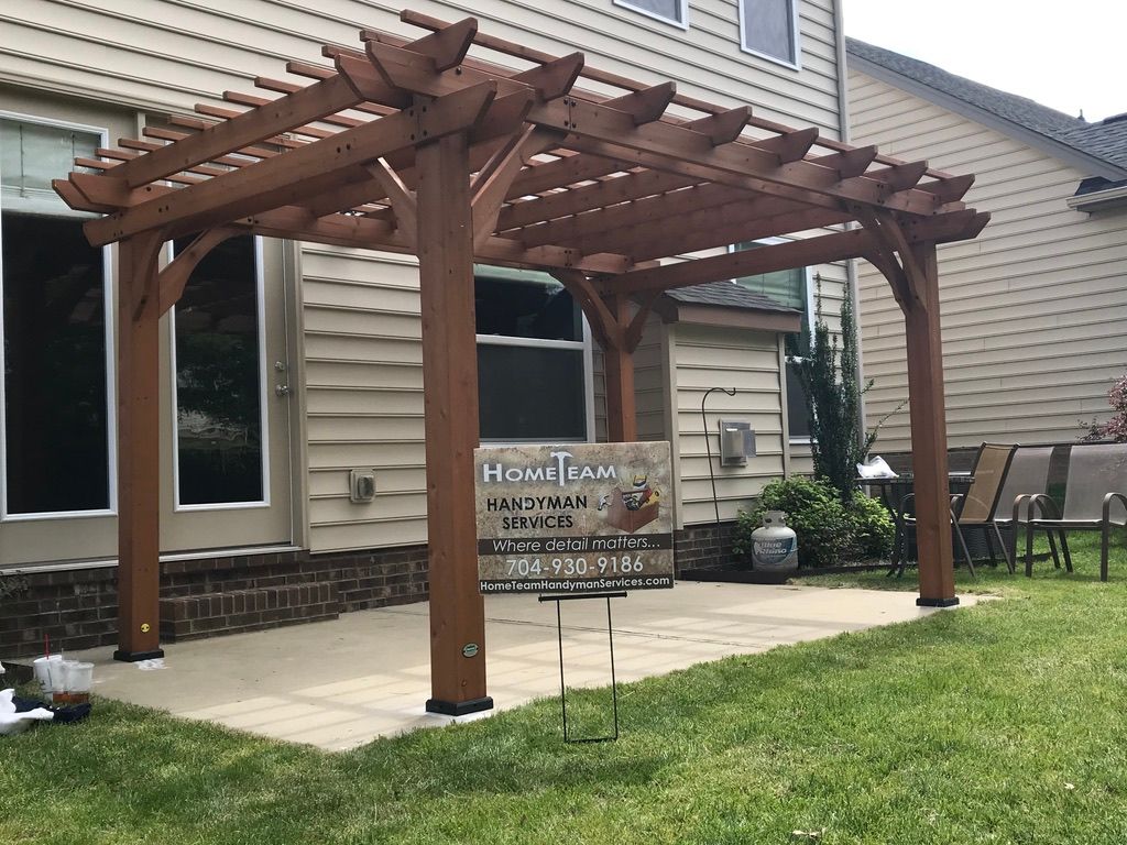Wooden pergola on a concrete patio in a backyard. The pergola is brown, and the house has beige siding.