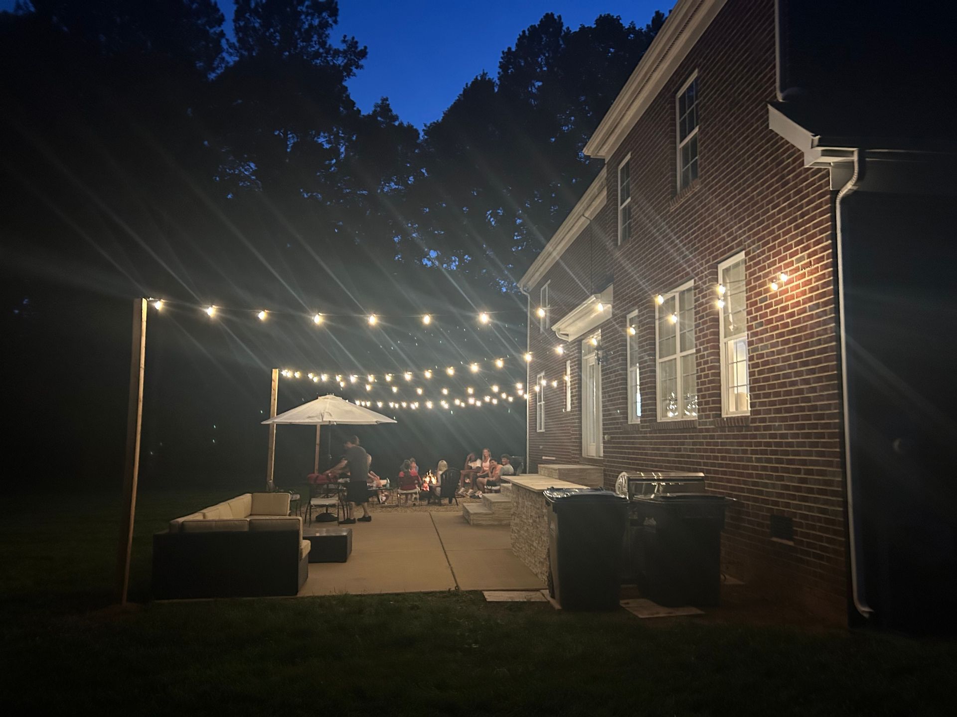 Nighttime outdoor patio with string lights. A group of people sits around a table under an umbrella. Brick house in background.