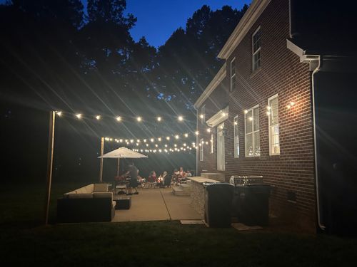 Patio party at night with string lights, a brick house, and people seated at tables under an umbrella.