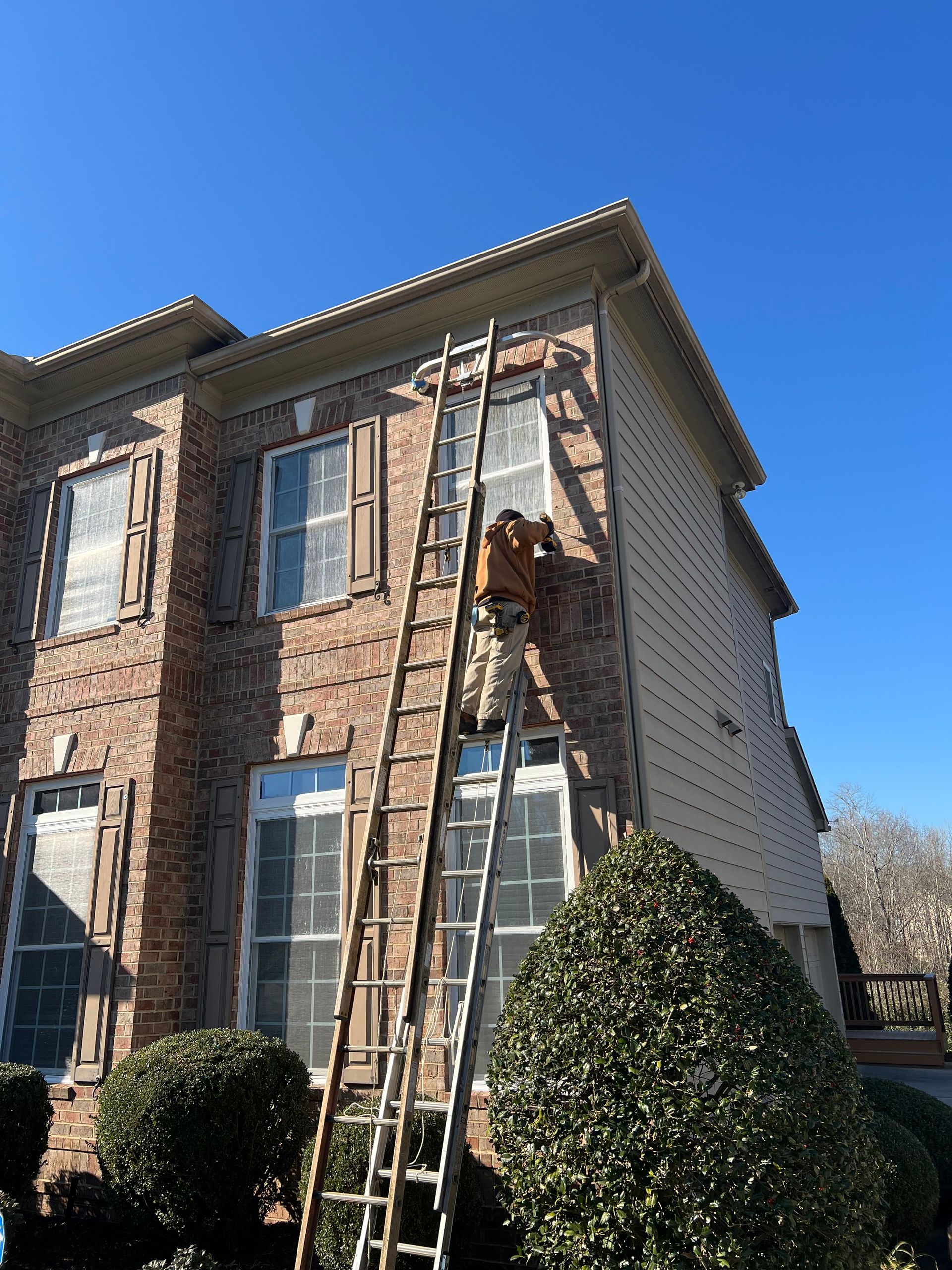 A person on a tall ladder is working on the side of a brick house under a clear, blue sky.