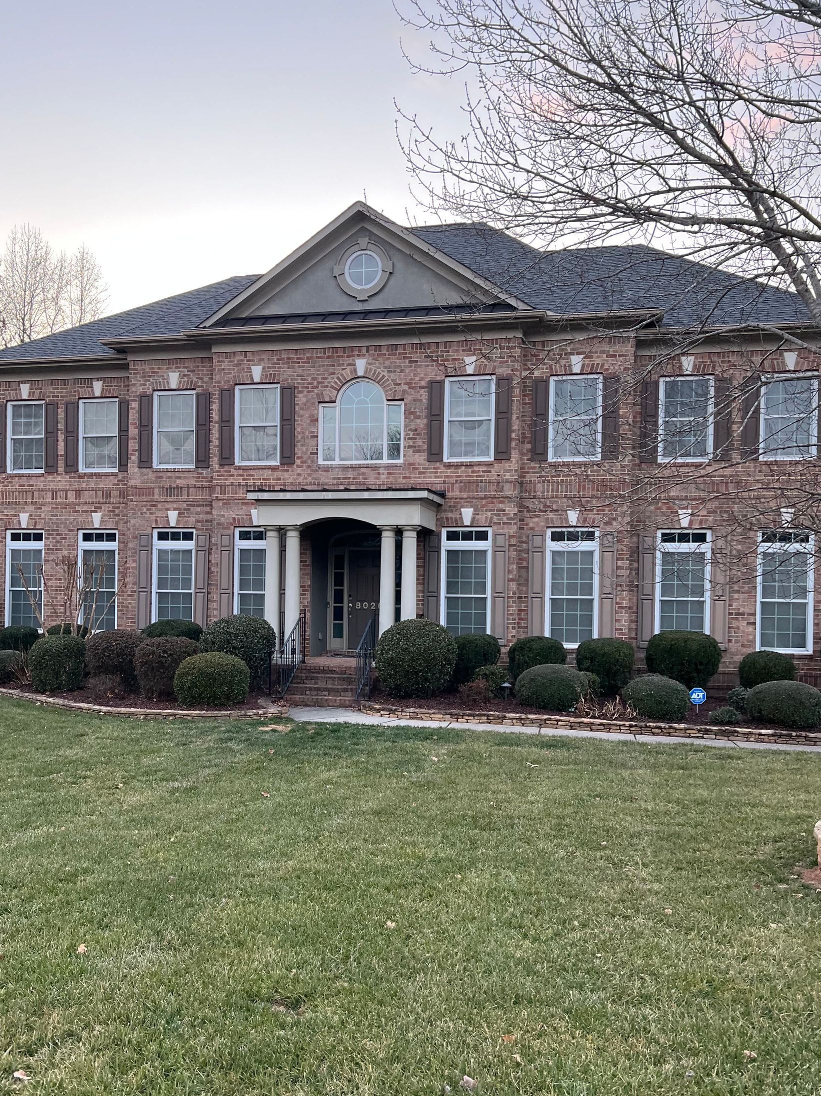 Large, two-story brick house with many windows and a columned front entrance. The lawn is green and a tree is visible on the right.