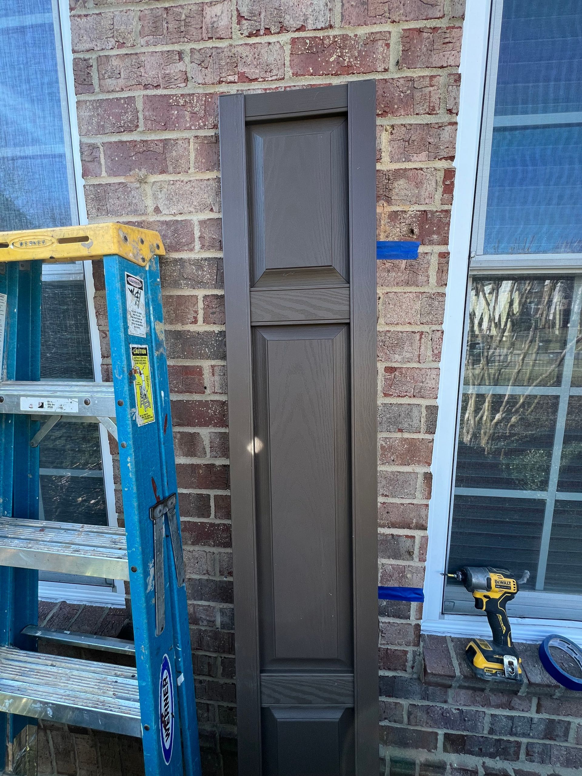 A brown shutter is propped against a brick wall next to a ladder and a window. A power drill and tape are on the windowsill.