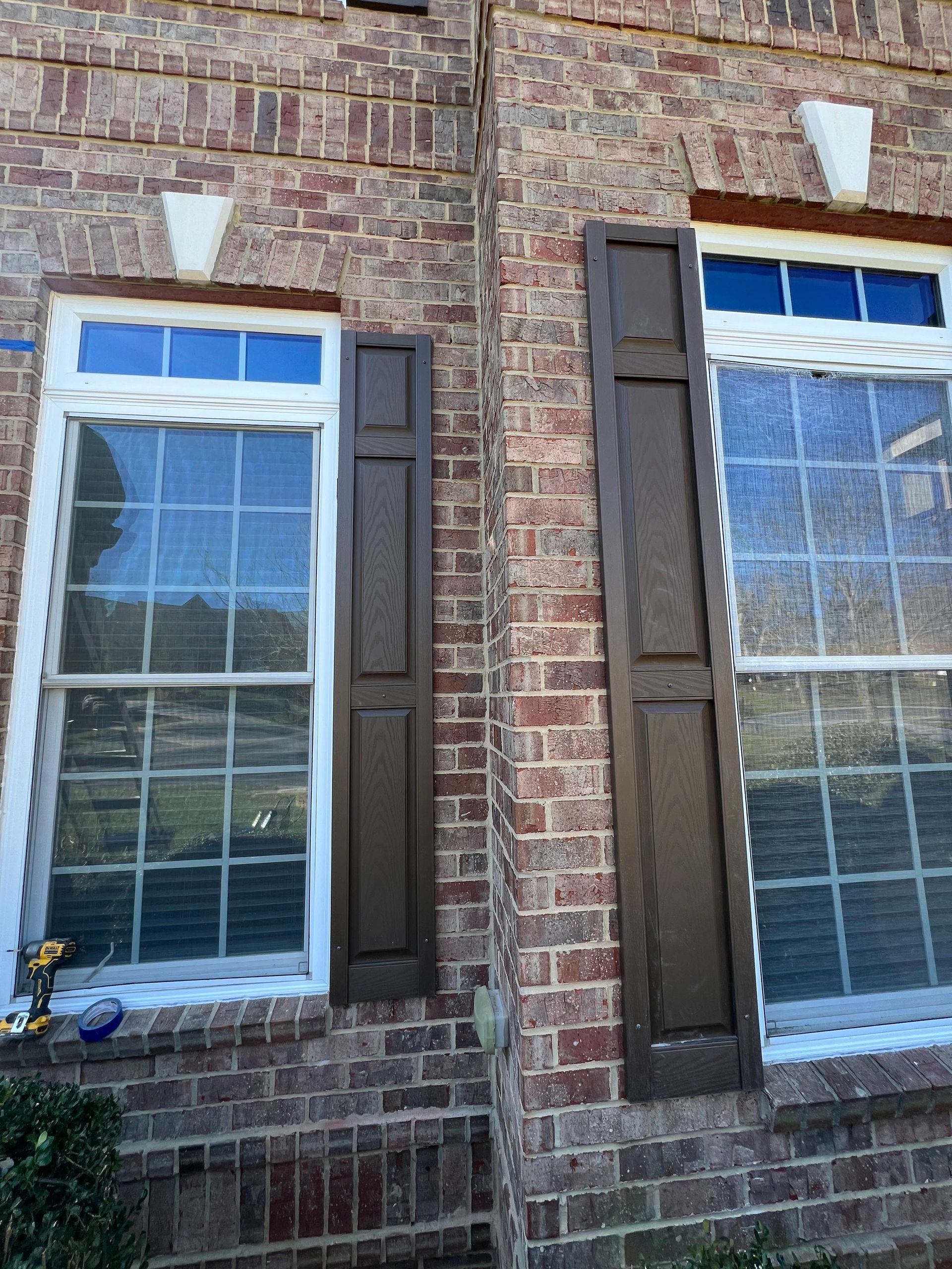 Exterior view of a brick house with two windows. Brown shutters are affixed to the wall between the windows. White trim surrounds the windows.
