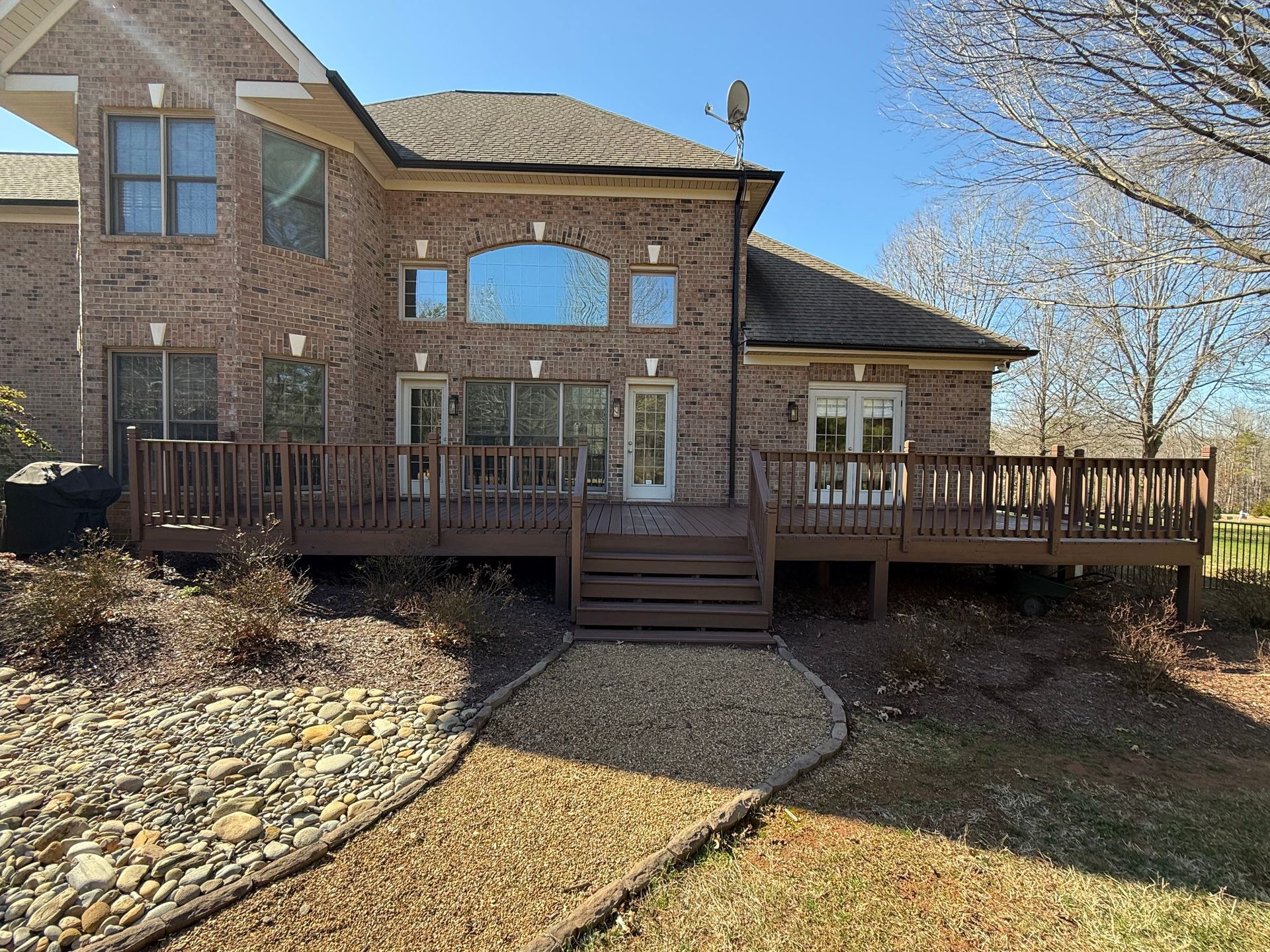 Back of a brick house with a brown deck, steps leading down to a gravel bed and grassy yard. Blue sky overhead.