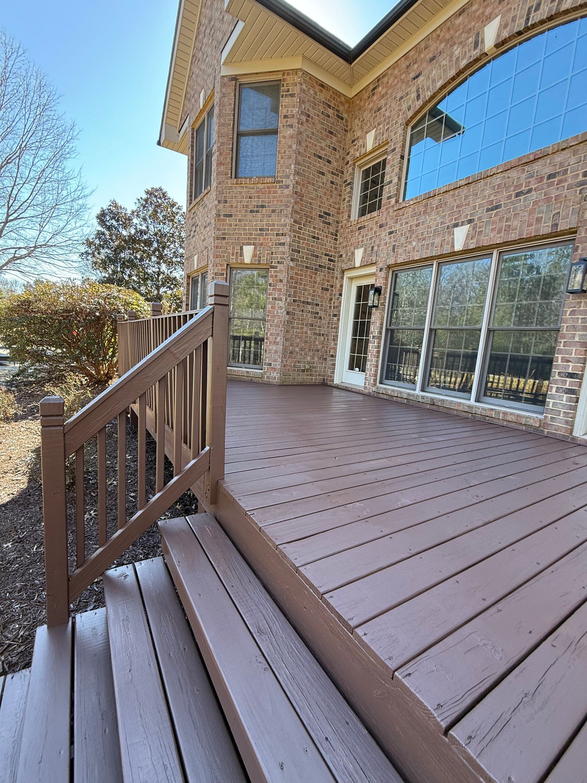 Brown wooden deck and stairs leading to a brick house with large windows under a sunny sky.