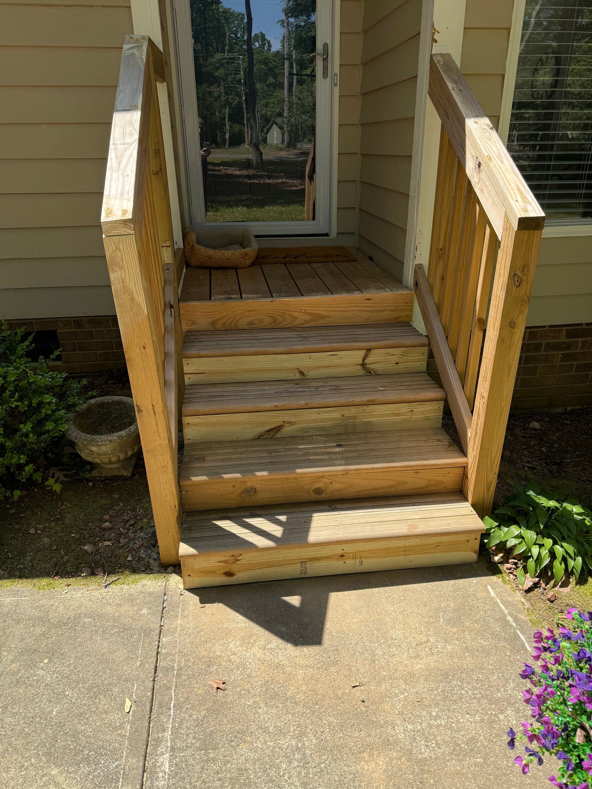Wooden exterior steps leading to a door, with a concrete walkway in front. The steps are weathered and have wooden railings.