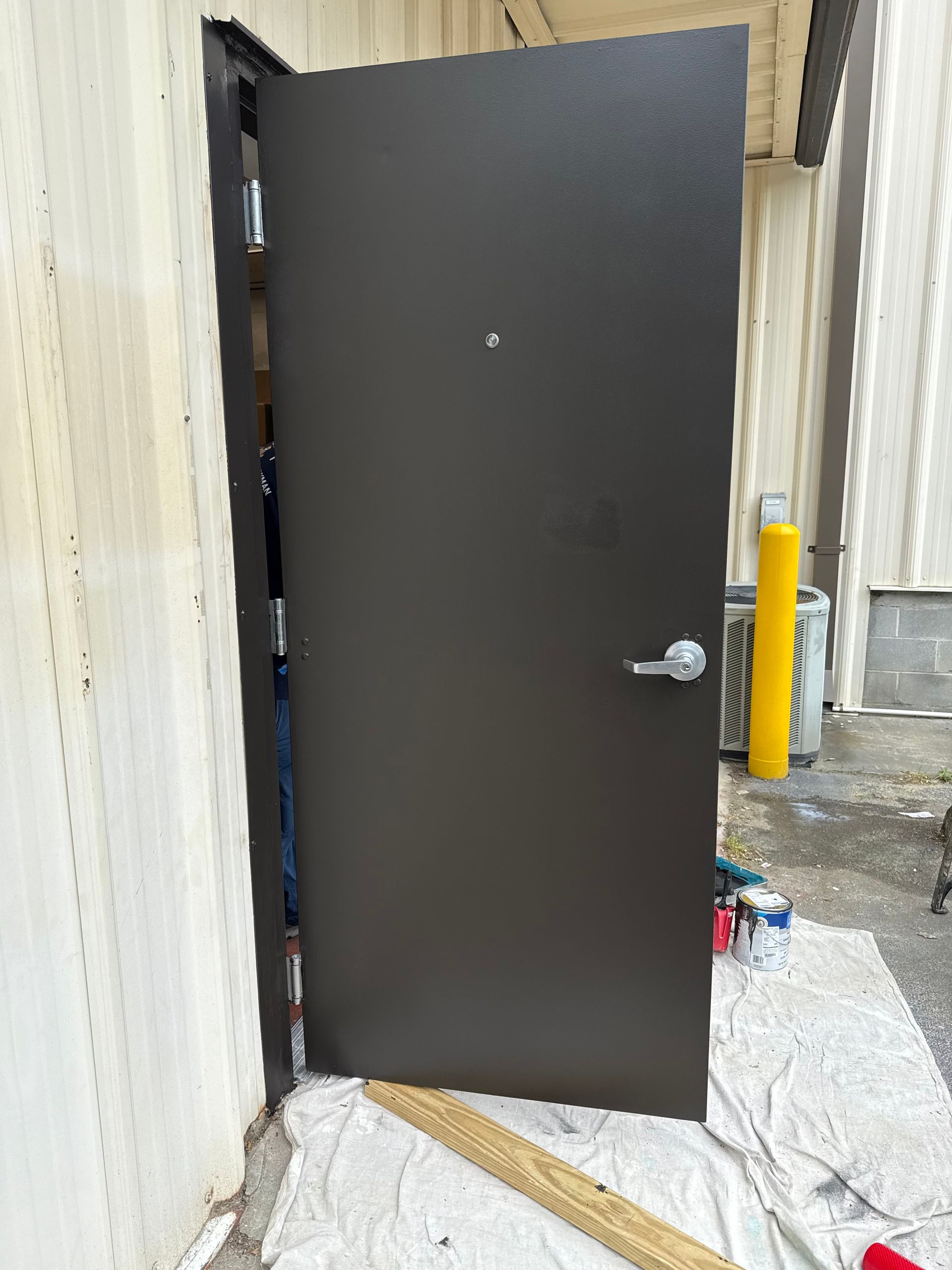 A dark brown metal door, partially open, is set in a white industrial building. A doorknob and peephole are visible.