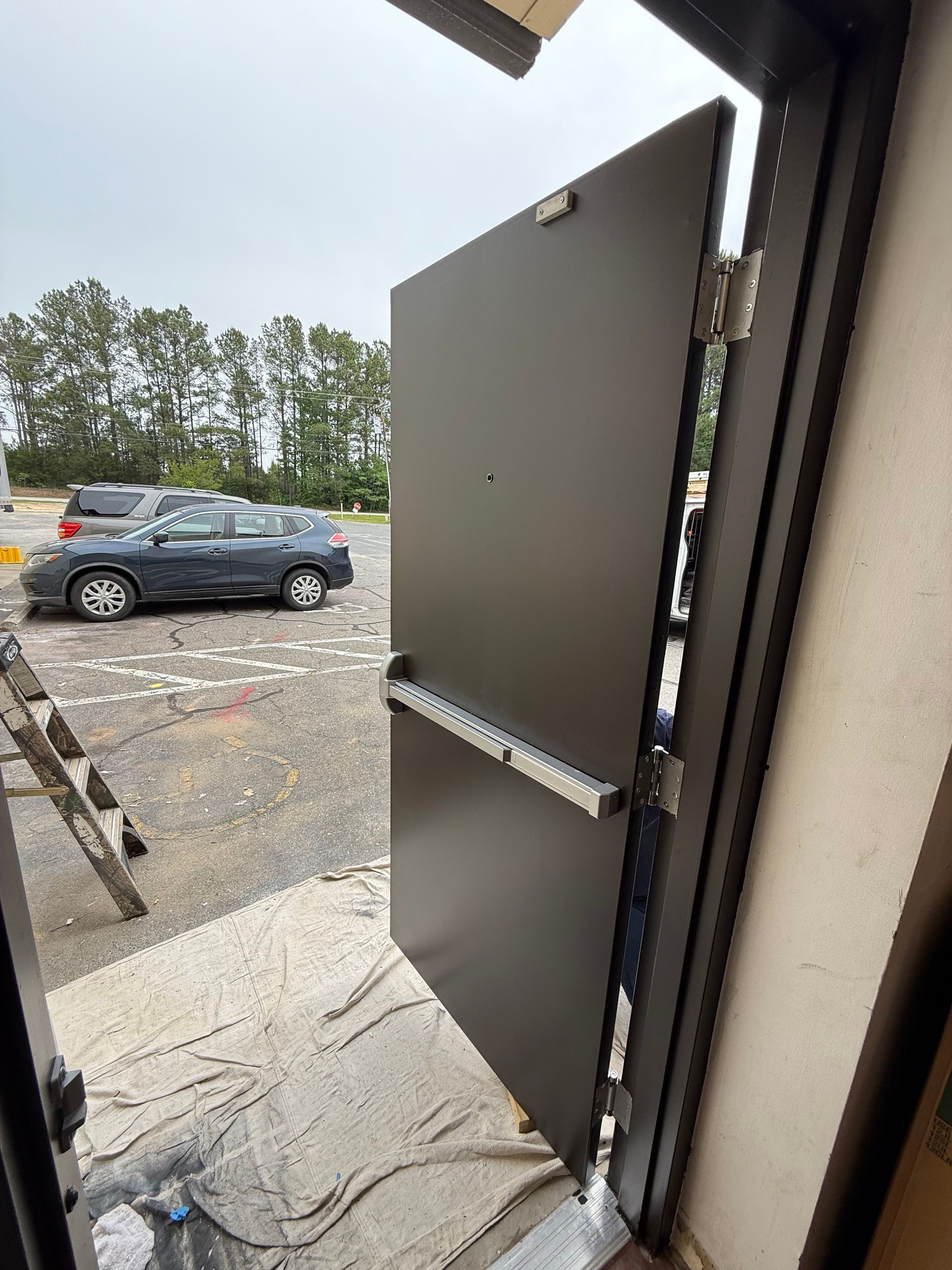 A partially open gray door with a silver push bar, leading to a parking lot with cars. The door has a keypad and is in an outdoor setting.
