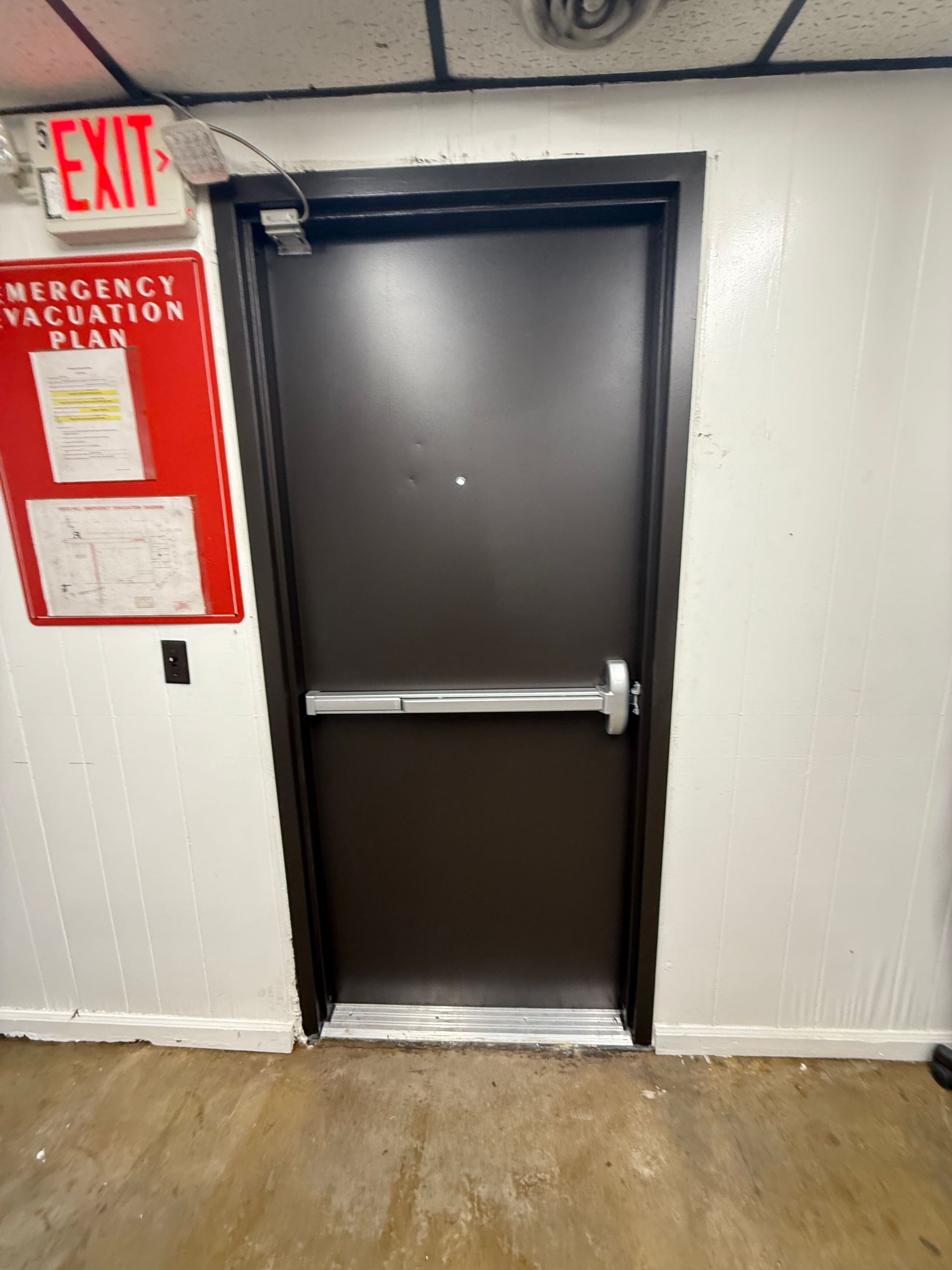 A brown exit door with a silver bar. Next to it is a red emergency evacuation plan and exit sign on a white wall.