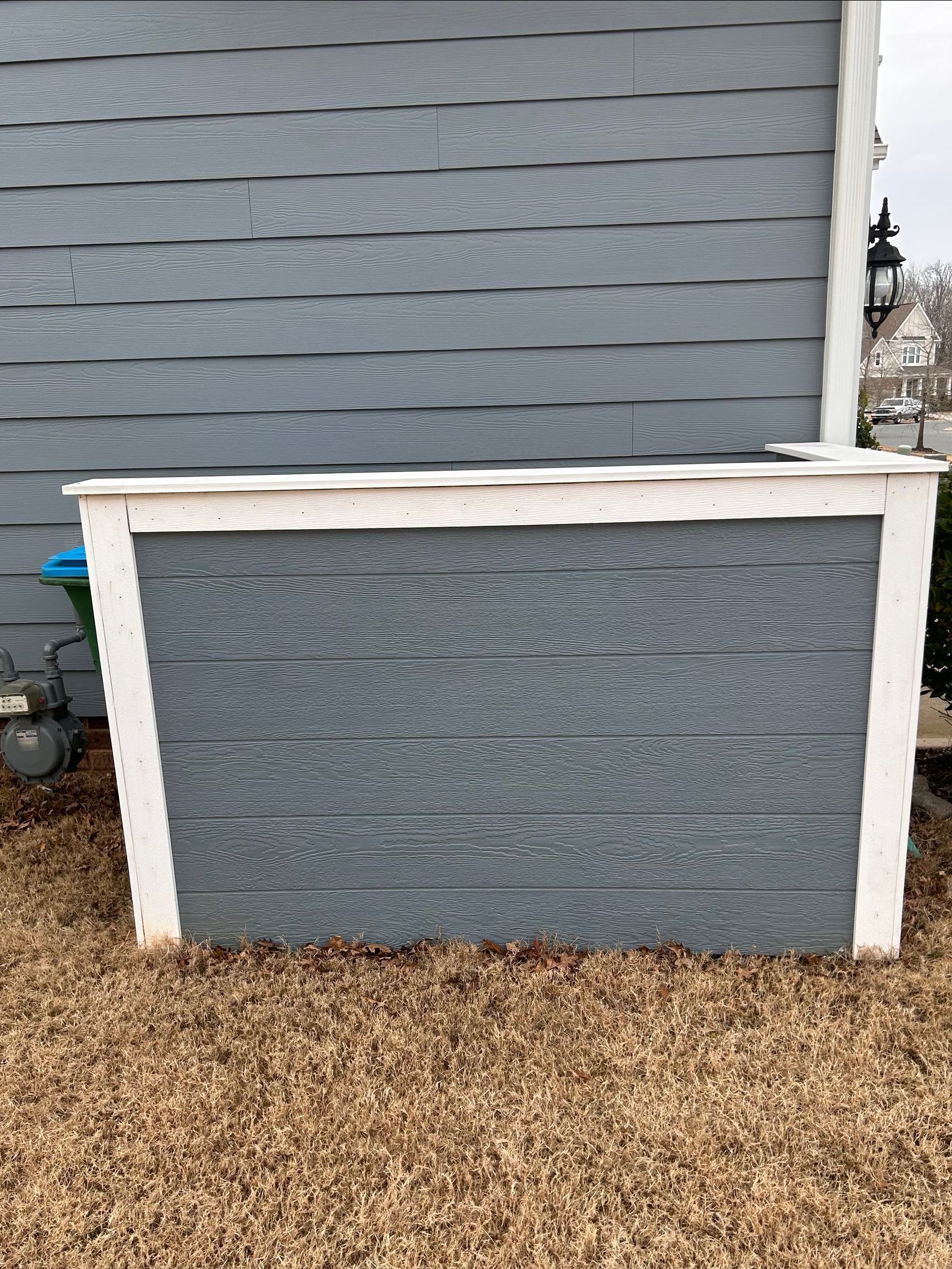 A wooden, rectangular box painted blue-gray with white trim sits outside on brown grass next to a blue house.