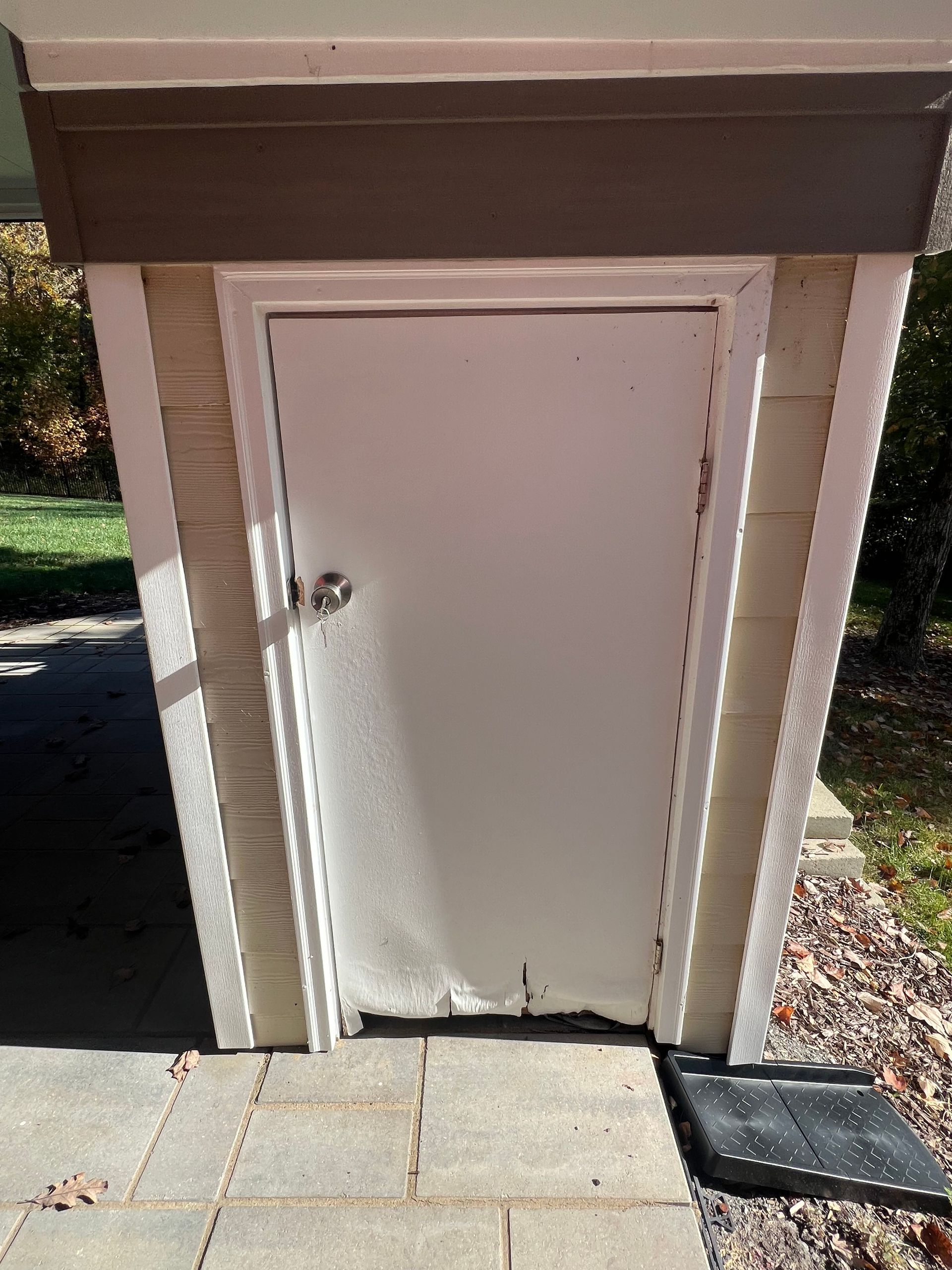White door with peeling paint, built into a beige-sided storage shed, set on stone pavers. A black mat is at the base of the door.