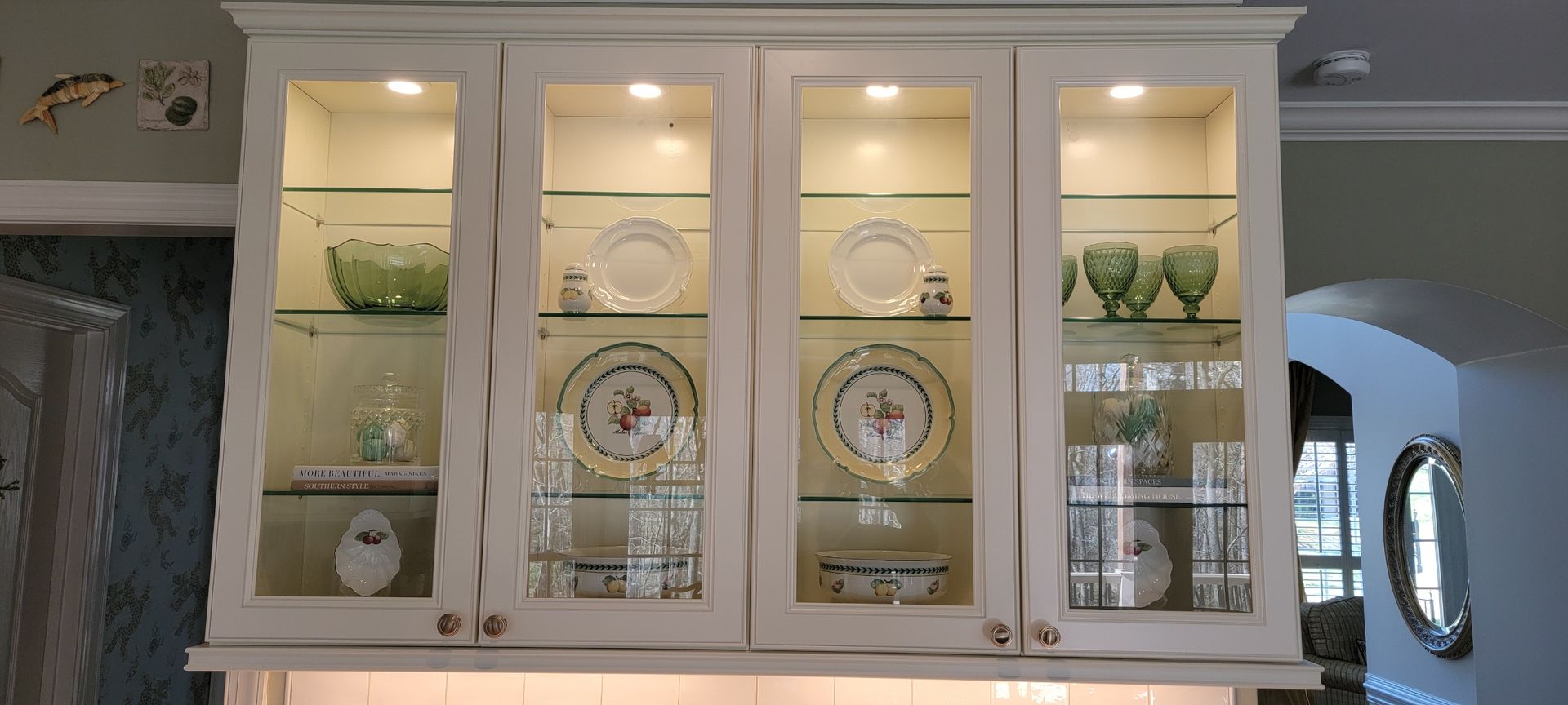 White kitchen cabinet with glass doors, illuminated inside, displaying dishes and glassware.