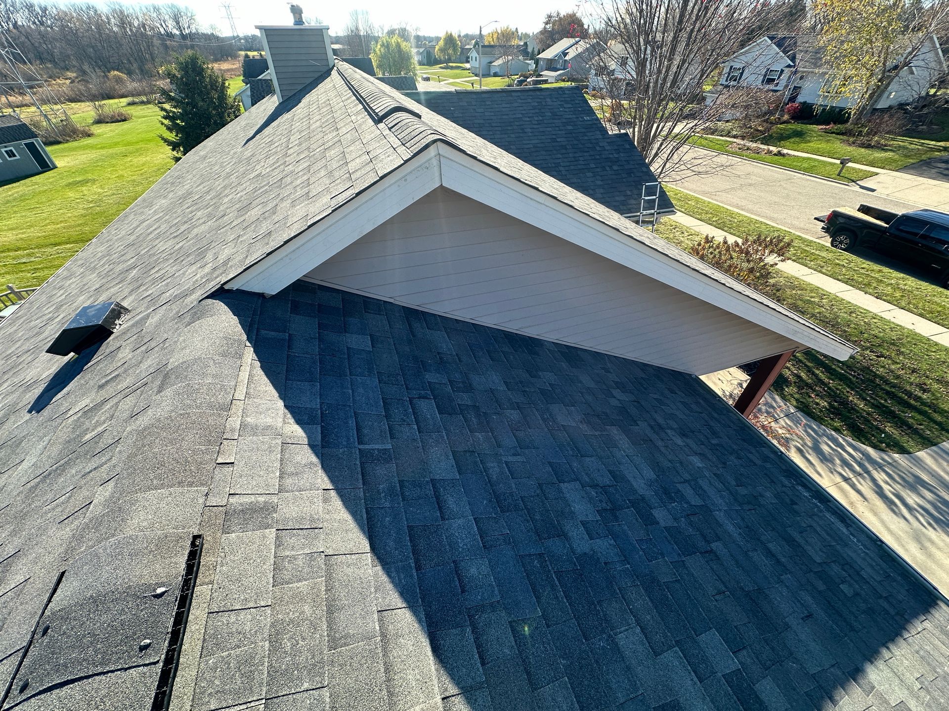 A house roof with dark gray shingles and a light beige gable against a backdrop of green grass and suburban homes.
