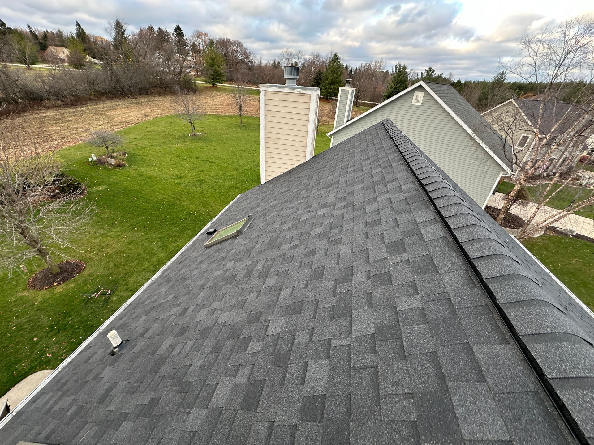 Dark gray shingled roof with a chimney and green grass, viewed from above on an overcast day.