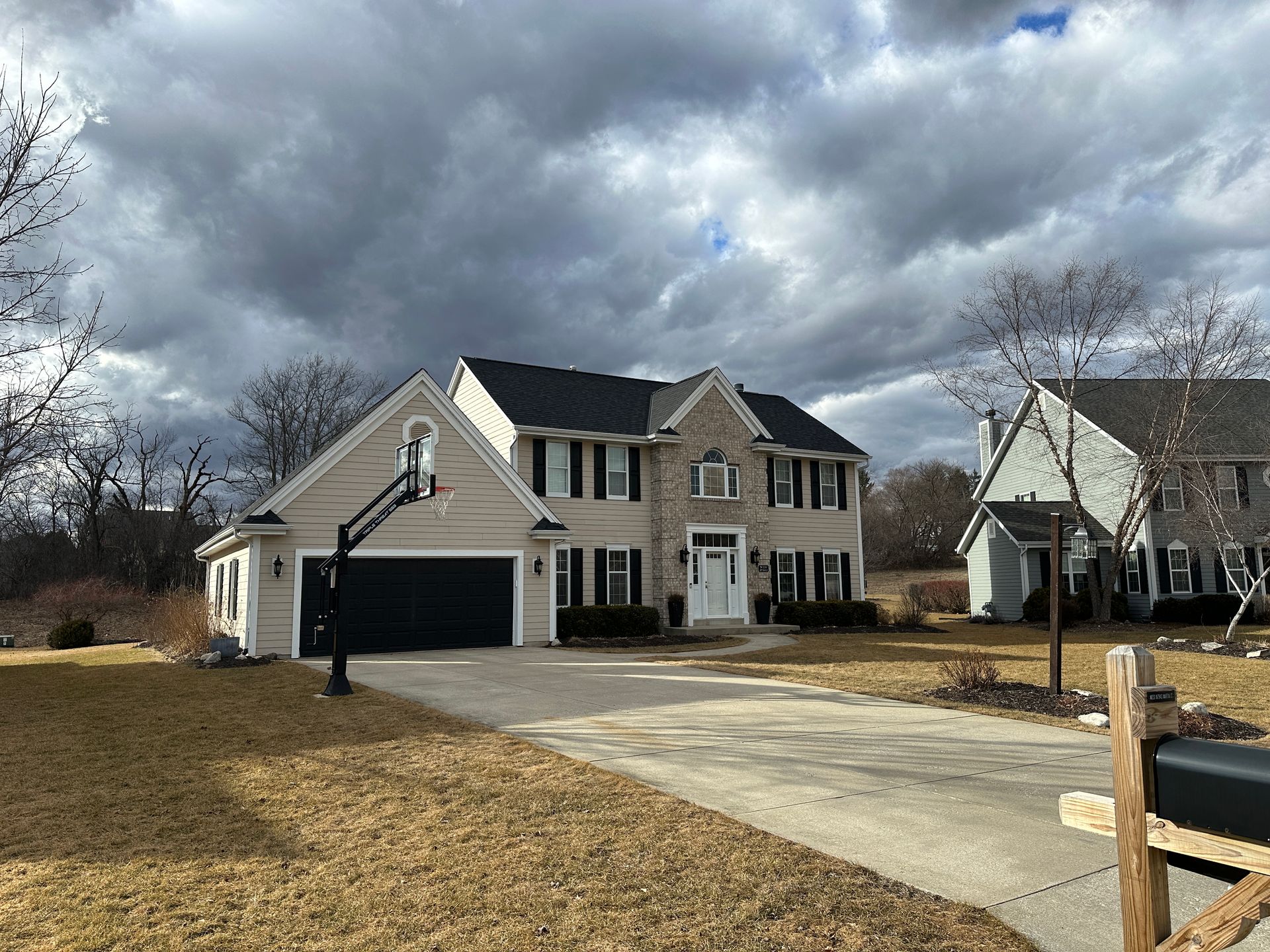 Two-story beige house with black shutters, driveway, basketball hoop, and cloudy sky.