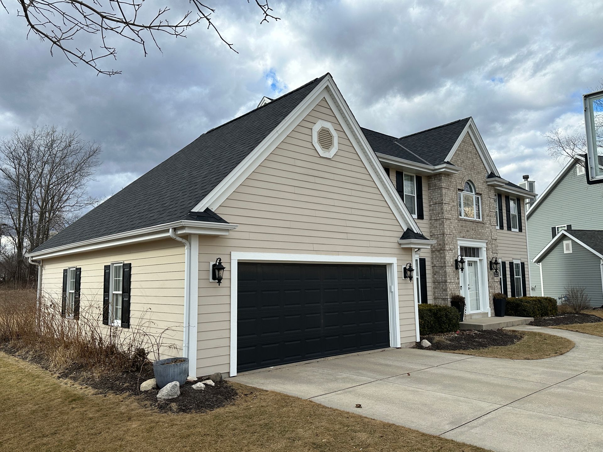 Beige house with black garage door, brick facade, and dark roof on a cloudy day.