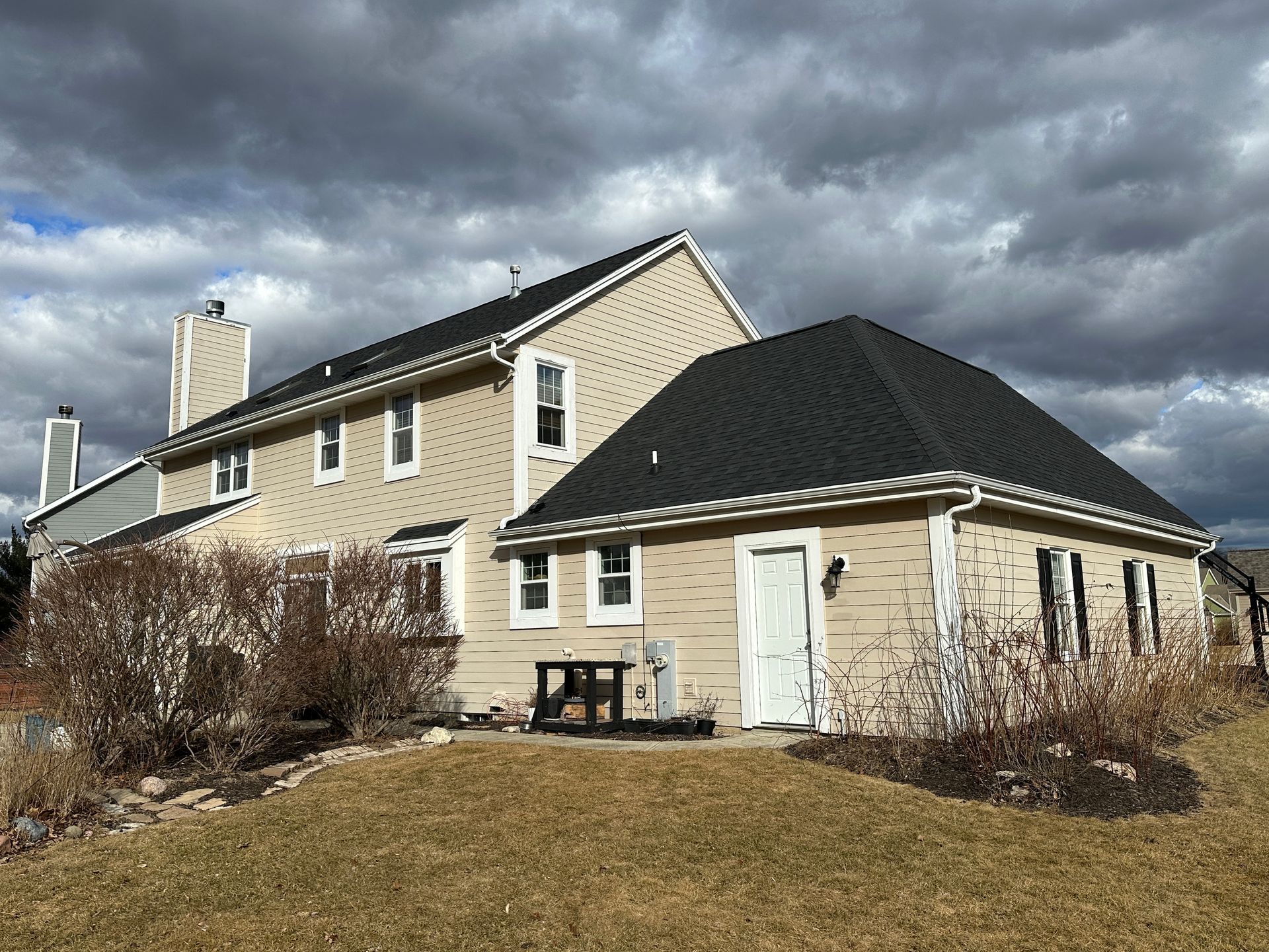 Beige house with a black roof, white trim, and a cloudy sky.