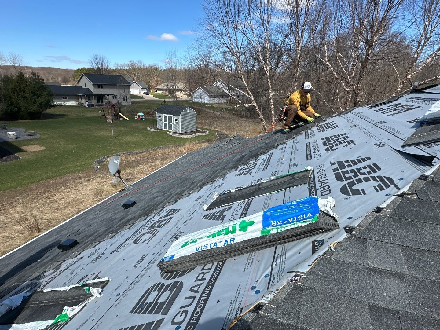Roofer installing shingles on a house roof, using safety harness. Overlaid with black protective underlayment.