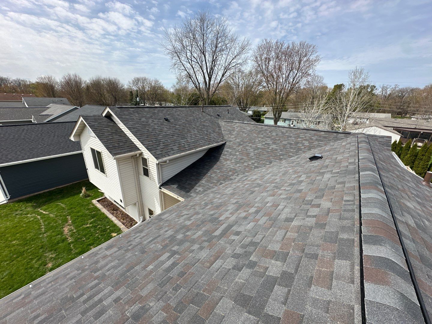 Overhead view of a house roof with gray asphalt shingles, surrounding trees, and blue sky.