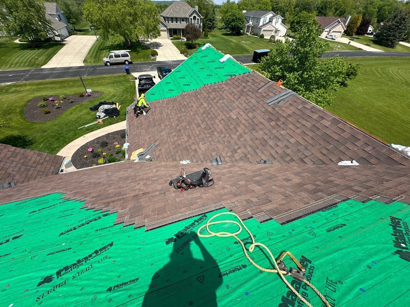 Roofers working on a brown shingle roof, green underlayment visible, sunny day.