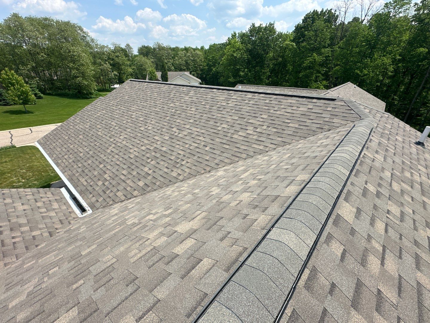 Asphalt shingle roof with gray and brown tones, on a house surrounded by green trees under a blue sky.