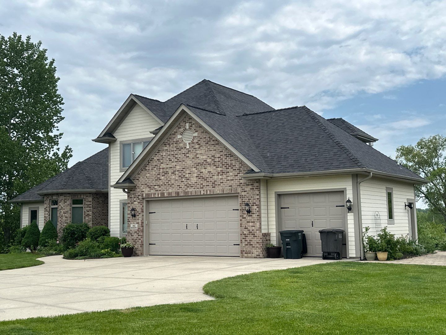 Two-story house with tan garage doors, light brick facade, and dark roof. Green grass and cloudy sky.