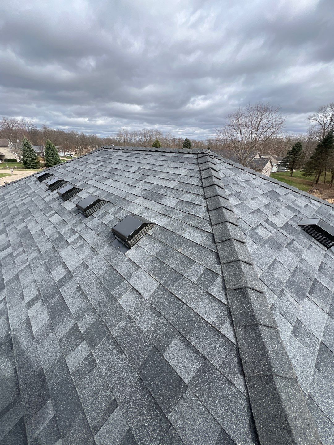 Close-up view of a shingled roof with several black vents under an overcast sky.