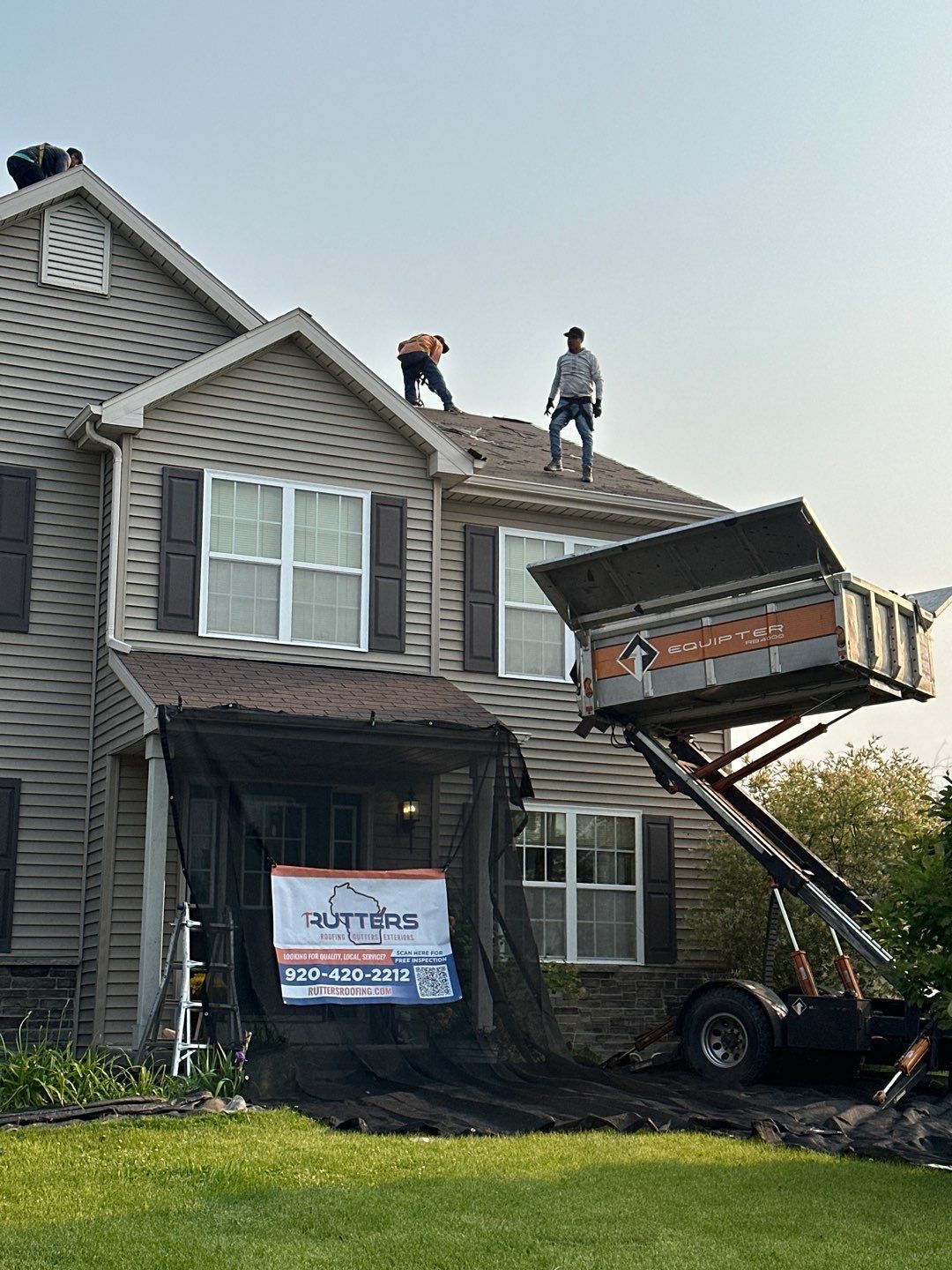 Two roofers on a house roof during repairs, with a lift truck and debris container.