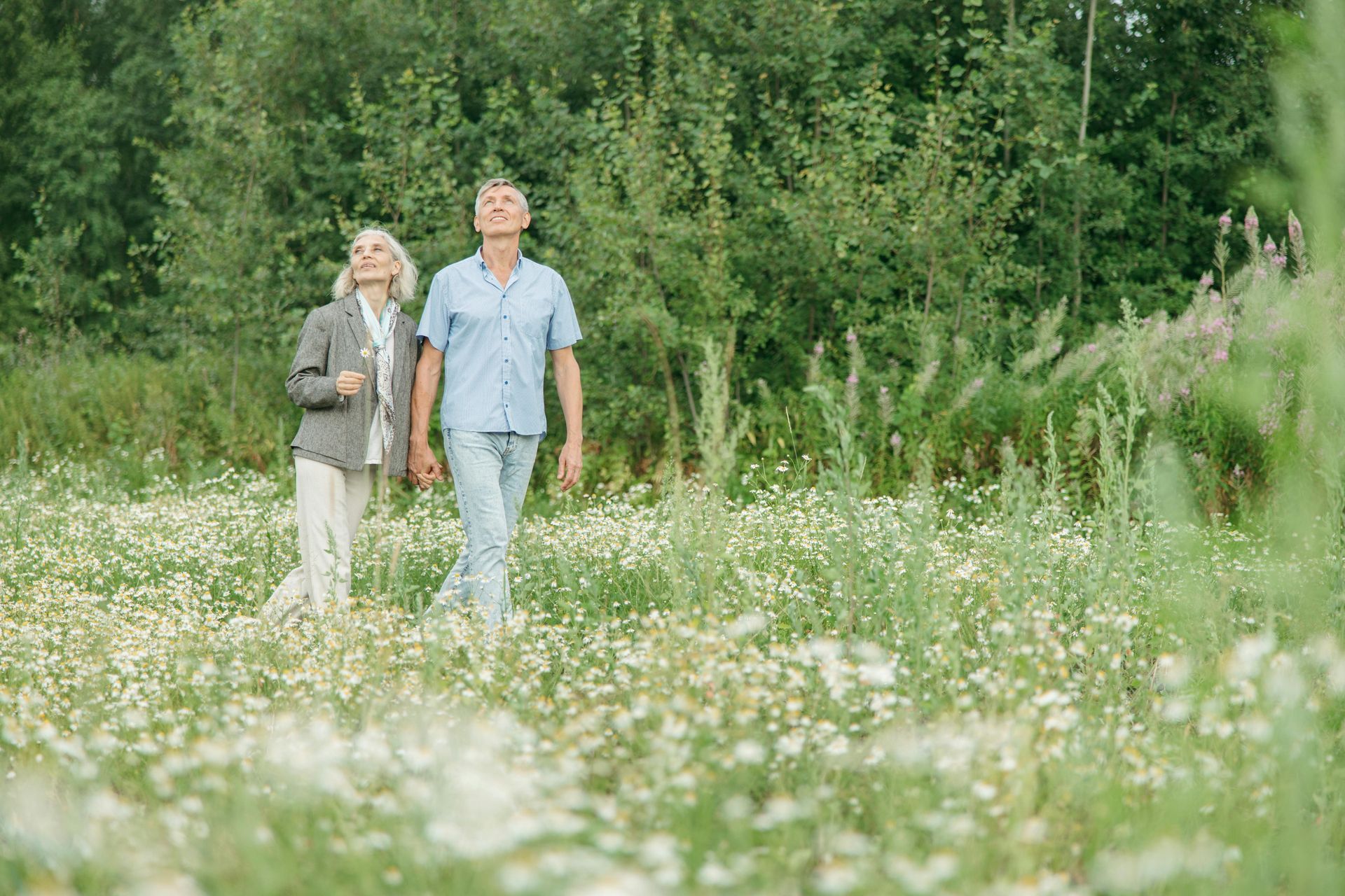 Couple walking through a field of wildflowers, looking upwards. Sunny day with green trees in the background.