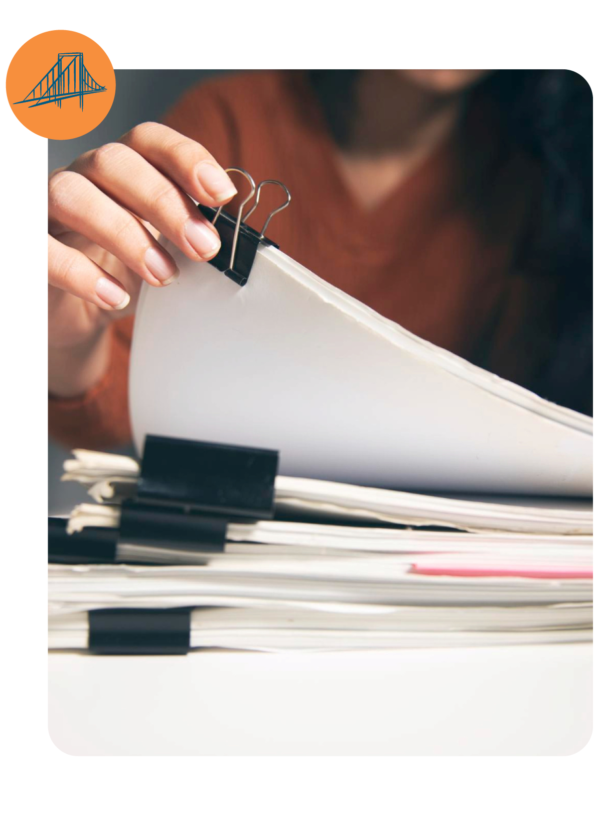 Hand holding a binder clip on stacked papers with a laptop in the background