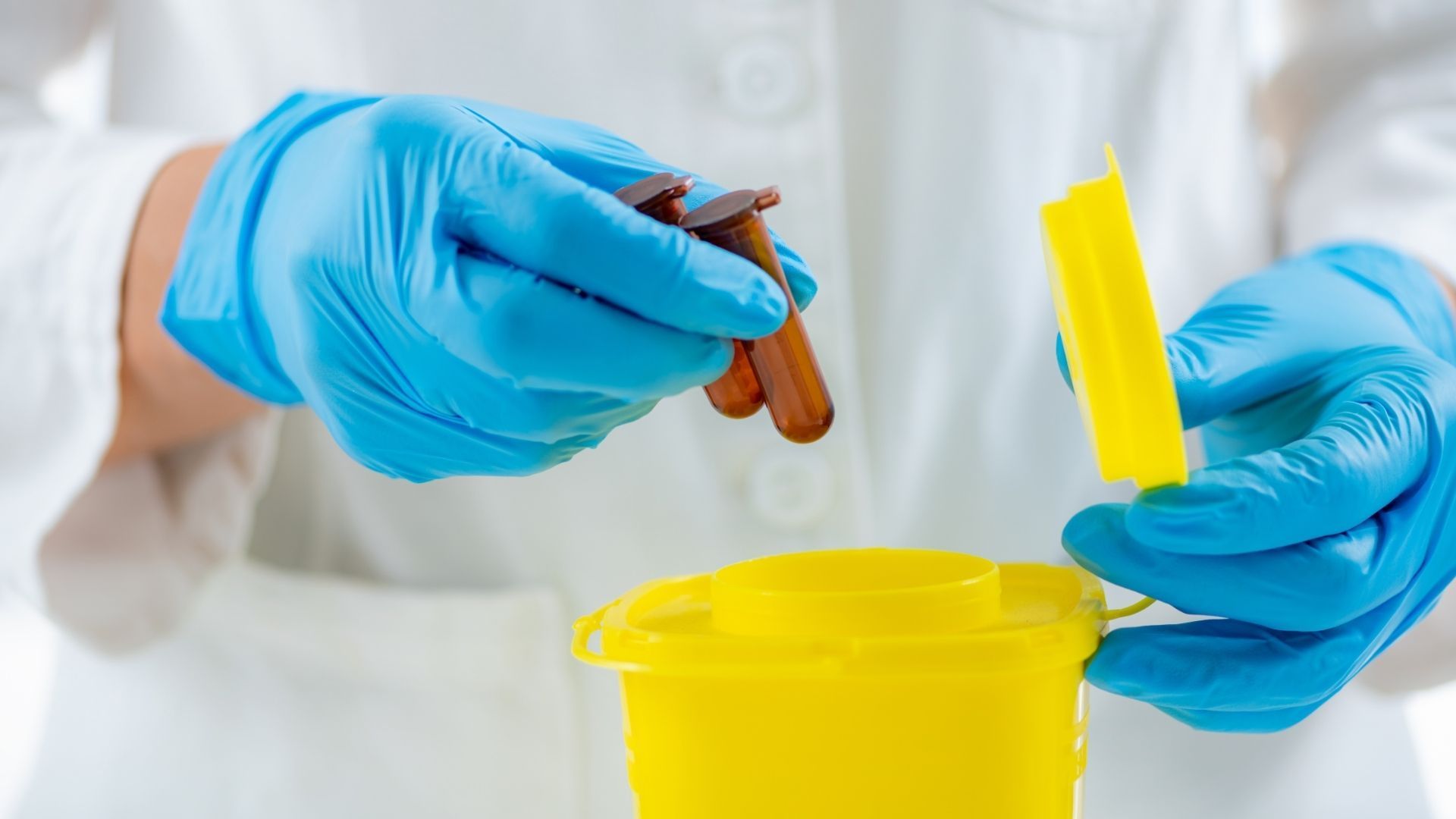 A person in a white lab coat and blue gloves places used syringes into a yellow hazardous waste container.