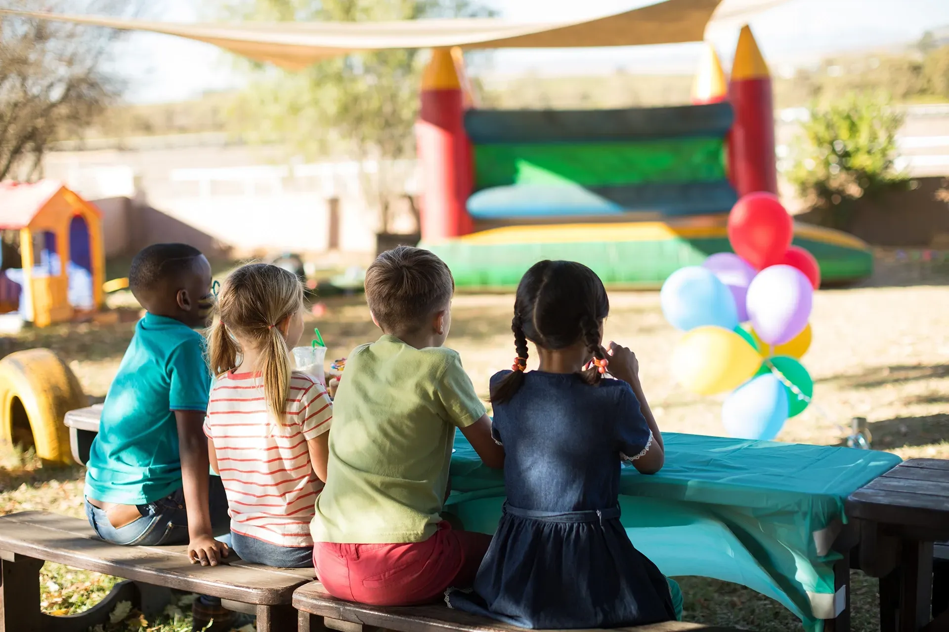 Eine Gruppe Kinder sitzt an einem Picknicktisch vor einem Hüpfburg.