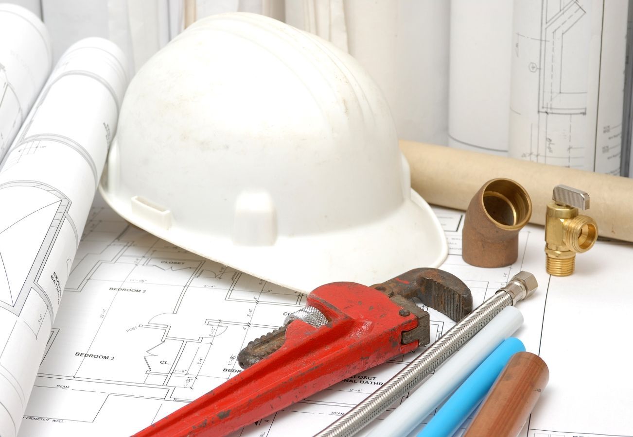 A hard hat sits on top of a blueprint next to a wrench and pipes