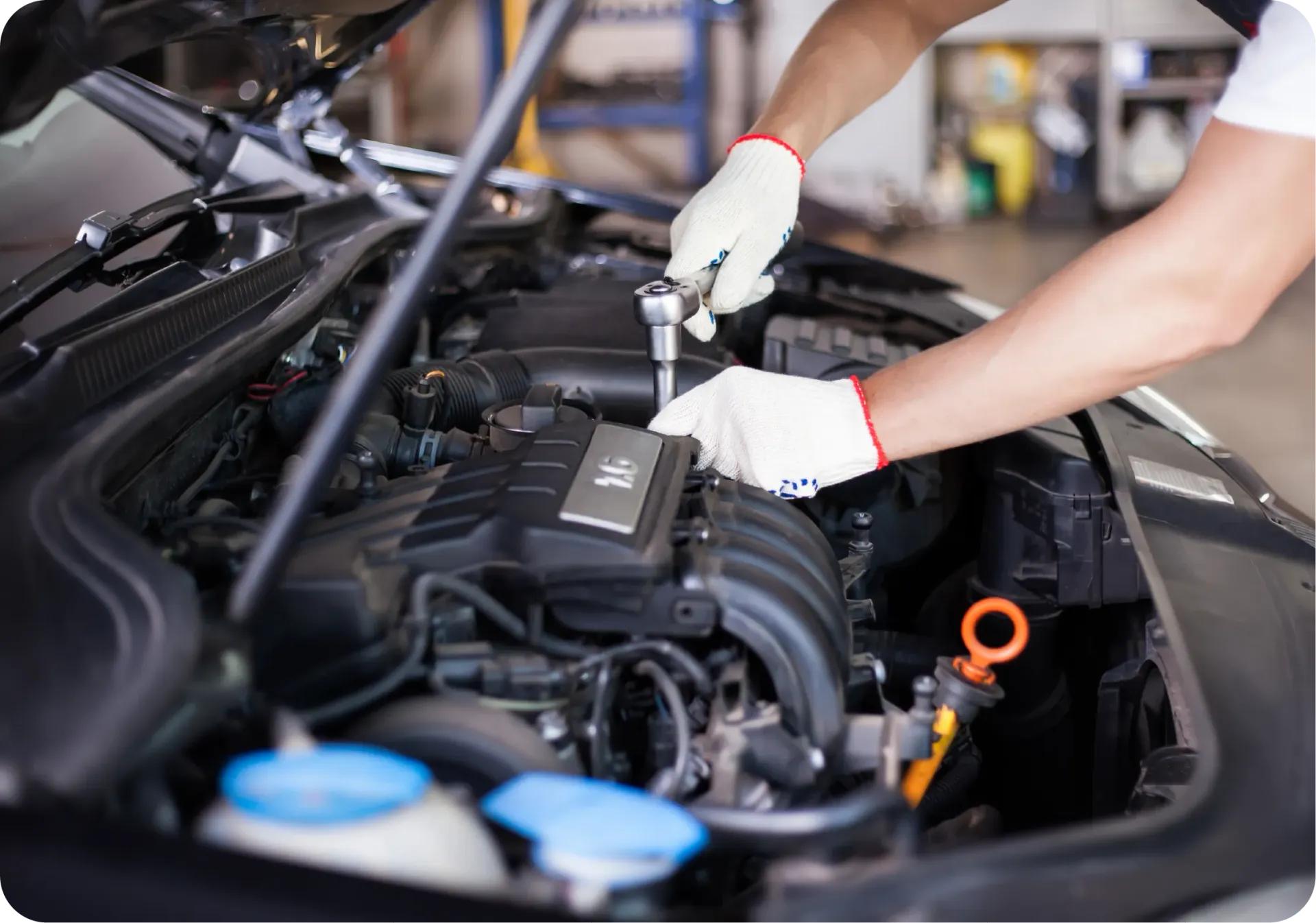 Mechanic wearing white gloves uses a ratchet tool to work on the engine of an open-hood vehicle in a workshop.