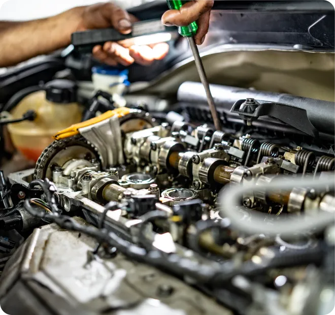 A close-up of a mechanic's hands using a screwdriver to work on an exposed car engine with a flashlight for illumination.