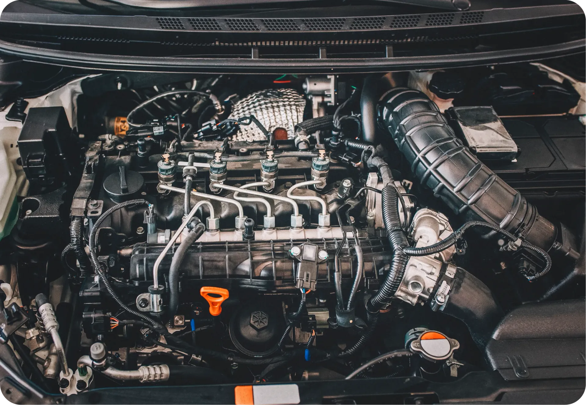 A top-down view of a modern car engine bay with various mechanical components, hoses, and a visible orange oil dipstick.