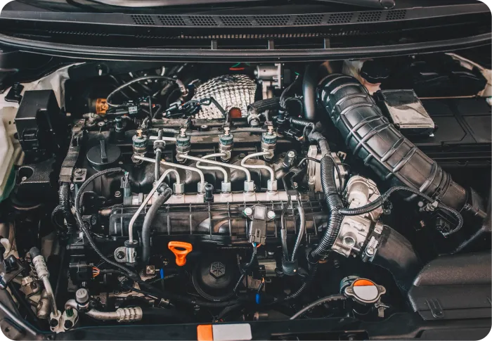 A top-down view of a modern car engine bay with various mechanical components, hoses, and a visible orange oil dipstick.