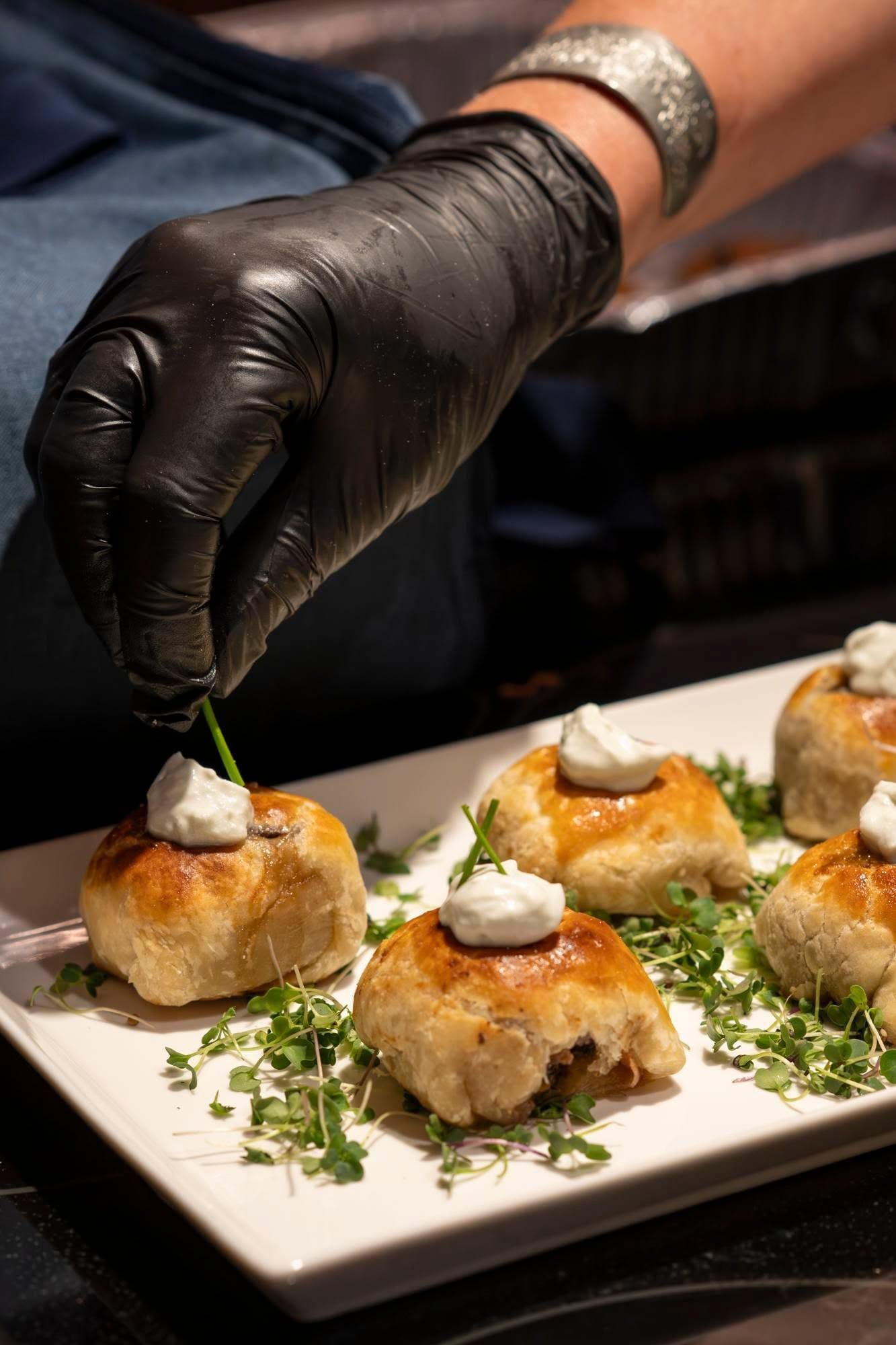 Gloved hand plating bite-sized appetizers on a white dish with herb garnish