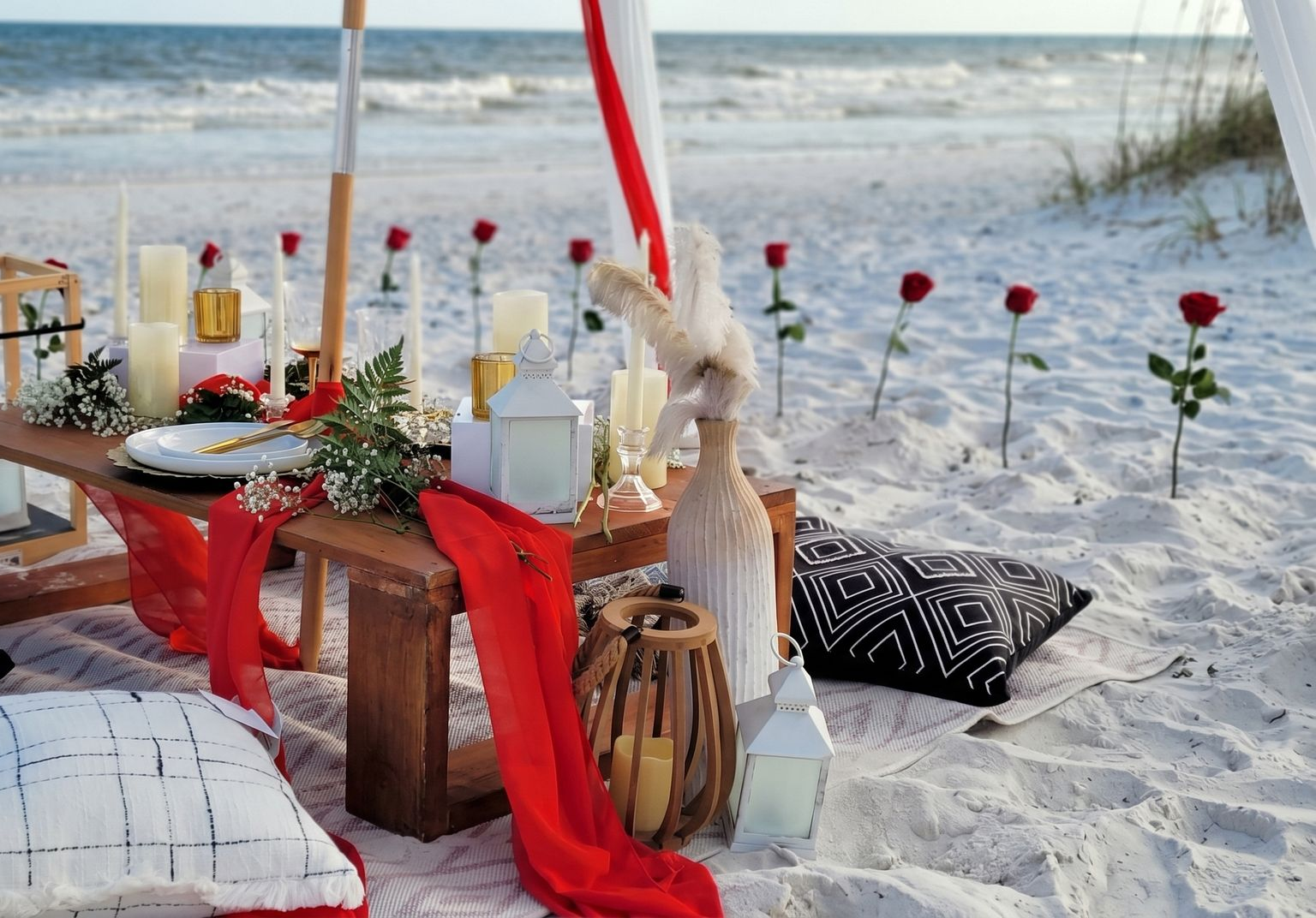 Beachside picnic table with red accents, candles, and white roses on the sand by the ocean